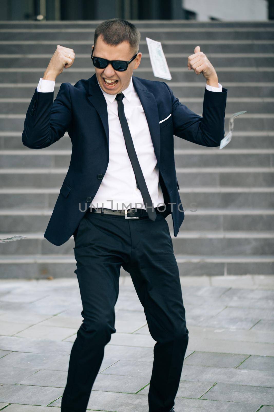 Portrait shot of cheerful rich handsome businessman in glasses and suit throws money. Happy good looking man throwing dollars. Money rain, falling dollars.