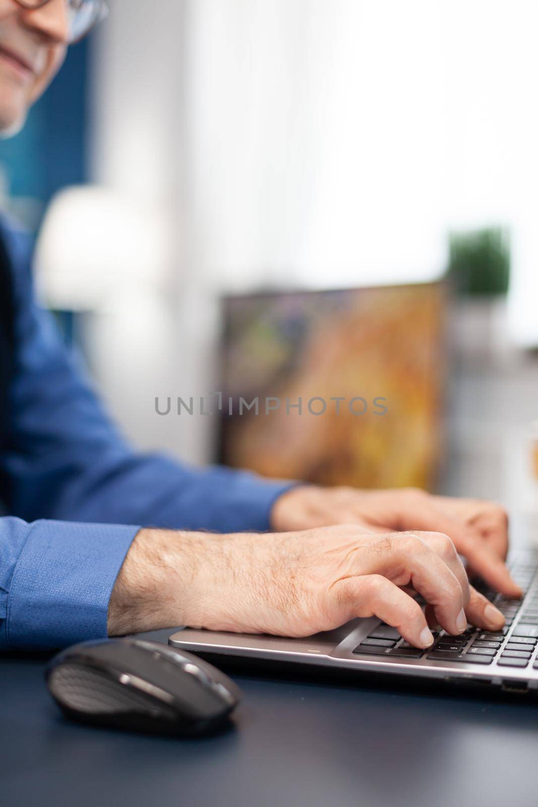 Close up of senior man hands typing on laptop keyboard. Elderly man entrepreneur in home workplace using portable computer sitting at desk while wife is holding tv remote