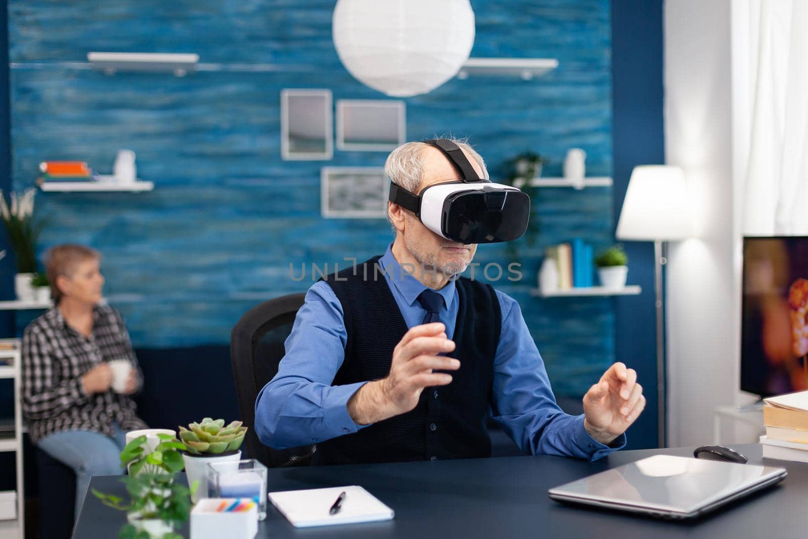Senior man wearing virtual reality headset doing hand gesturing. Elderly man pensioner in living room sitting at desk with futuristic goggles for augmented cyberspace wife watching tv.