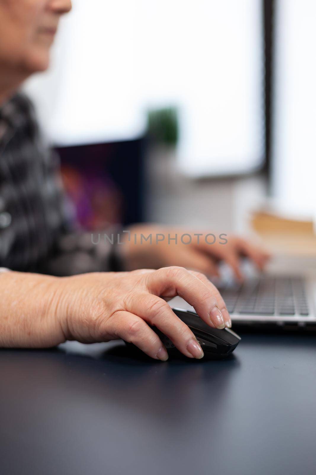 Close up of old entrepreneur lady using mouse in home office. Elderly woman in home living room using moder technoloy laptop for communication sitting at desk indoors.