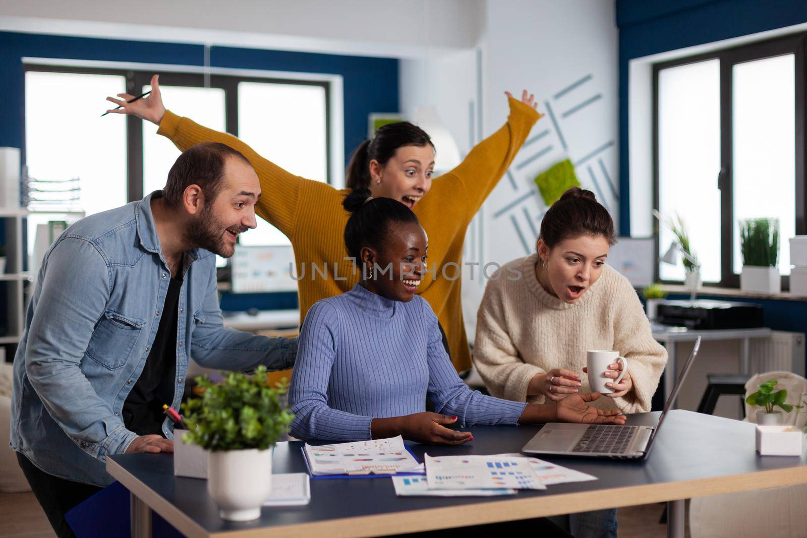 Diverse startup coworkers entrepreneurs celebrating triump with hands up lin modern office. Multiethnic business team with laptop and papers excited about project.