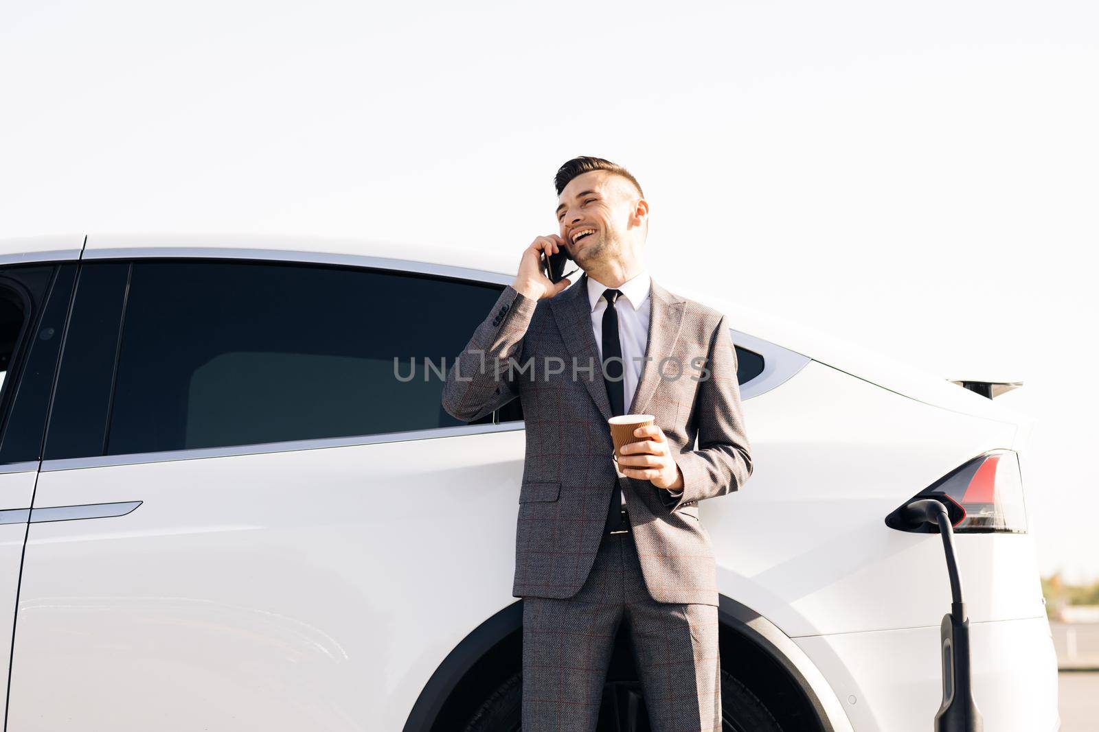 Businessman is talking on his Smart Phone while his electric car is charging on background. Side view of white luxury car. Environmentally conscious male charging electric vehicle by uflypro