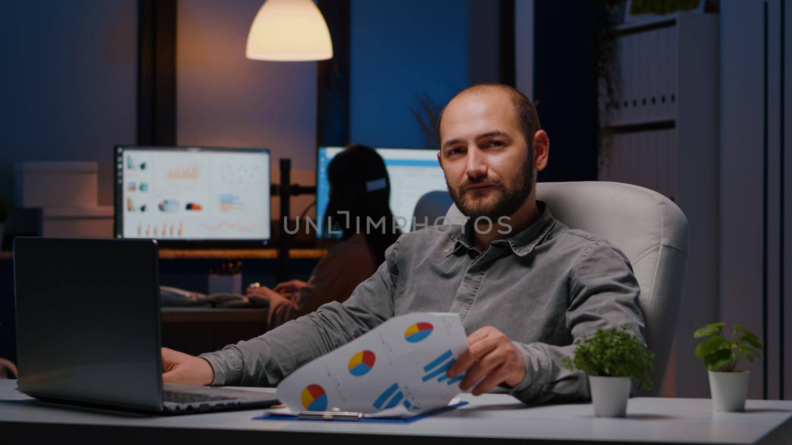 Portrait of smiling businessman sitting at desk table analysing financial statistics on clipboard. Manager working at management strategy before company deadline late at night in company offfice