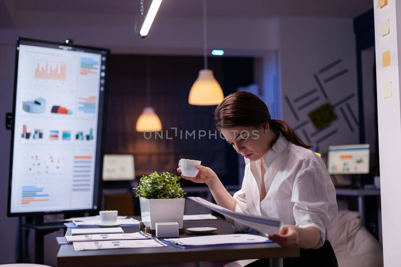 Workaholic businesswoman looking at reports presentation on monitor comparing with financial chart on paperwork. Focused manager working in business company meeting office room late at night