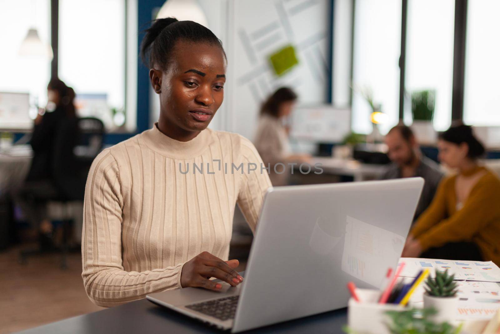Black manager reading tasks on laptop and typing sitting at desk in busy start up office while diverse team analyzes statistics data in background. Multiethnic group talking about project