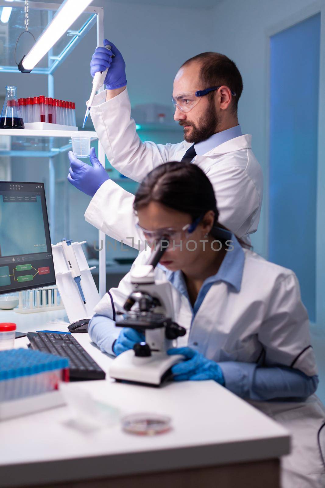 Scientist examining blood sample looking under microscope while chemist doctor using micropipette for pharmaceutical expertise. Team of medical researchers working to discovering cure against disease