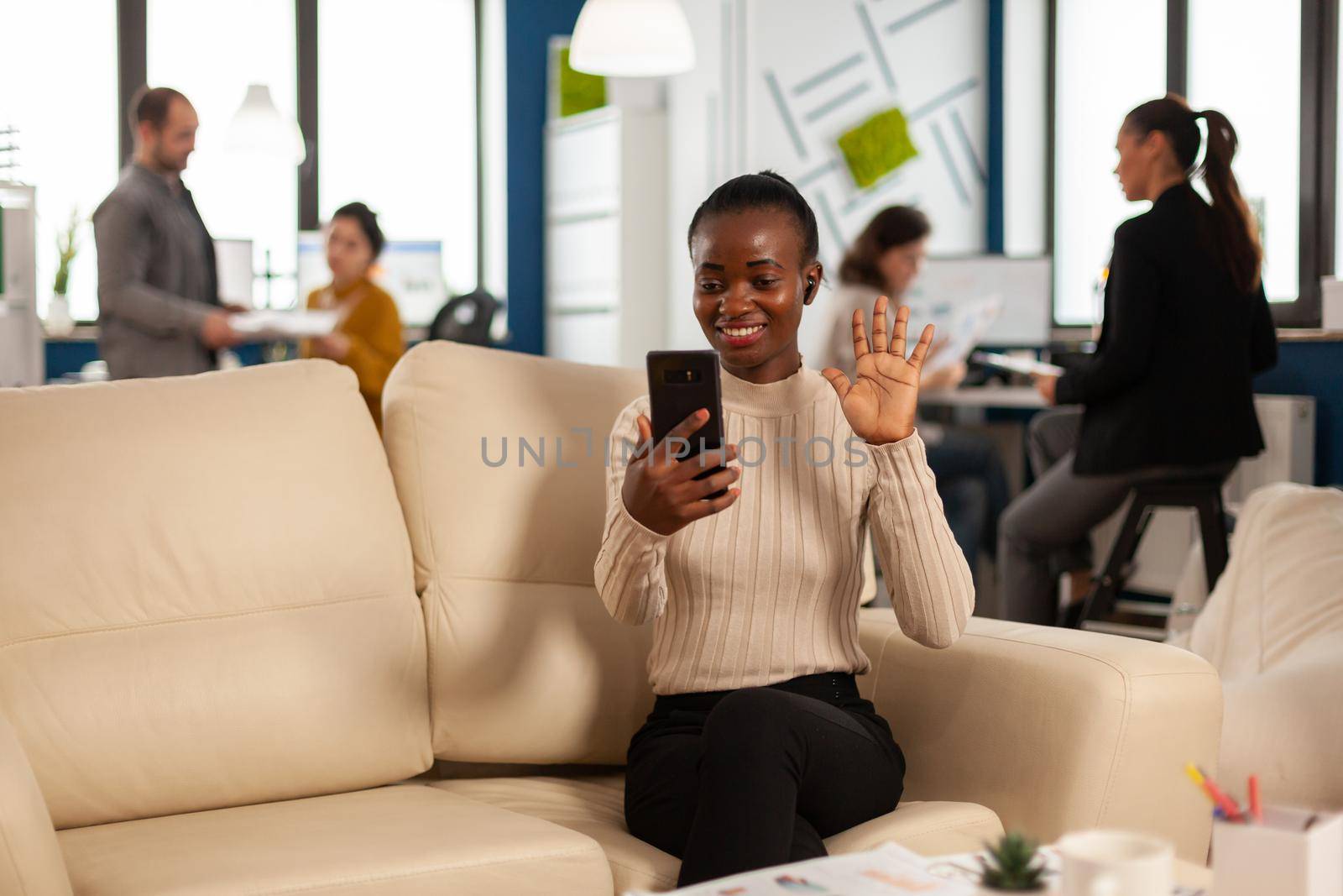 Black woman waving at camera explaining financial reports to remote manager on video call holding smartphone using headphones sitting on couch. Diverse coworkers planning financial project