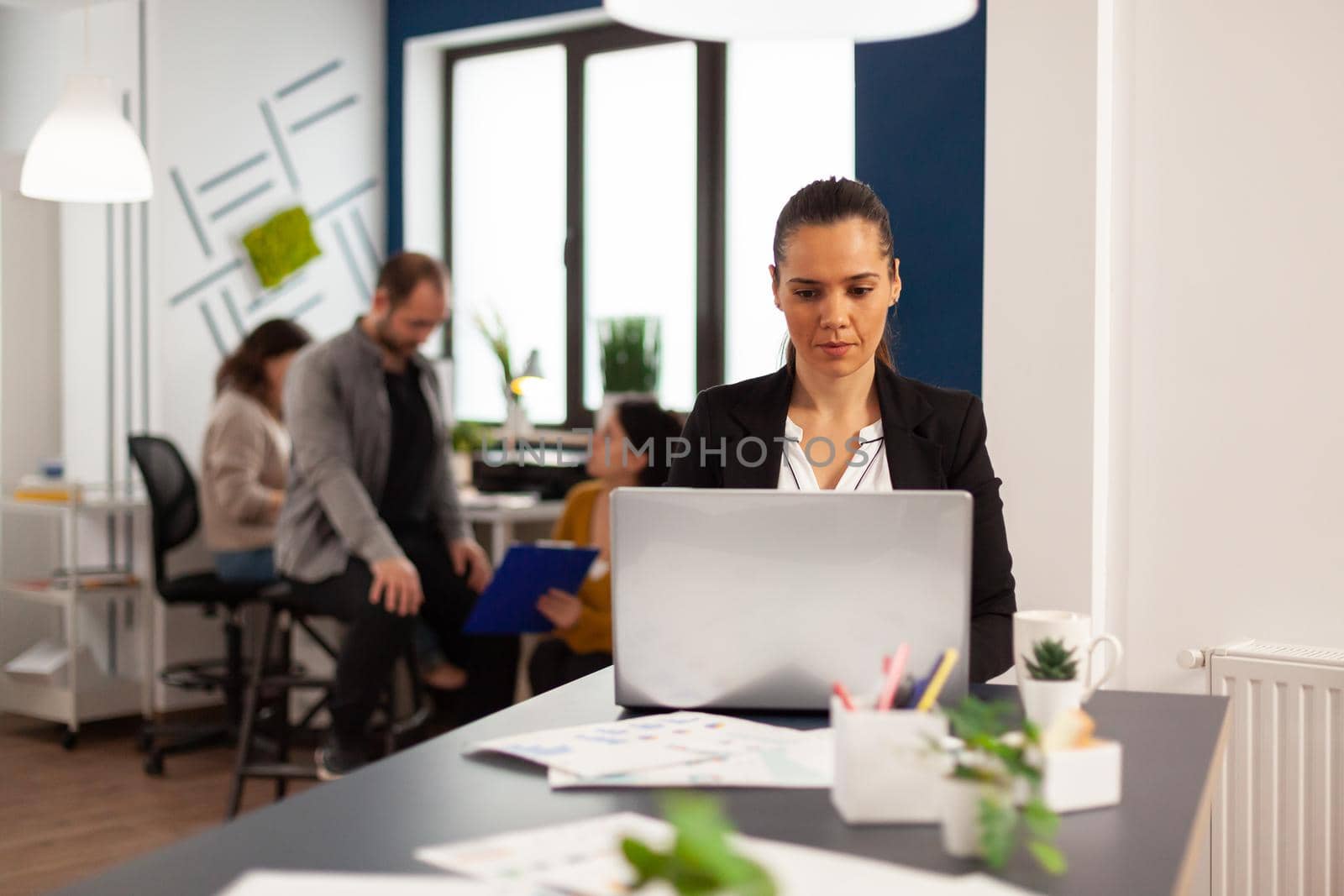 Happy hispanic lady typing on laptop sitting at desk in start up business office drinking coffee while diverse colleagues working in background. Multiethnic coworkers planning new financial project.