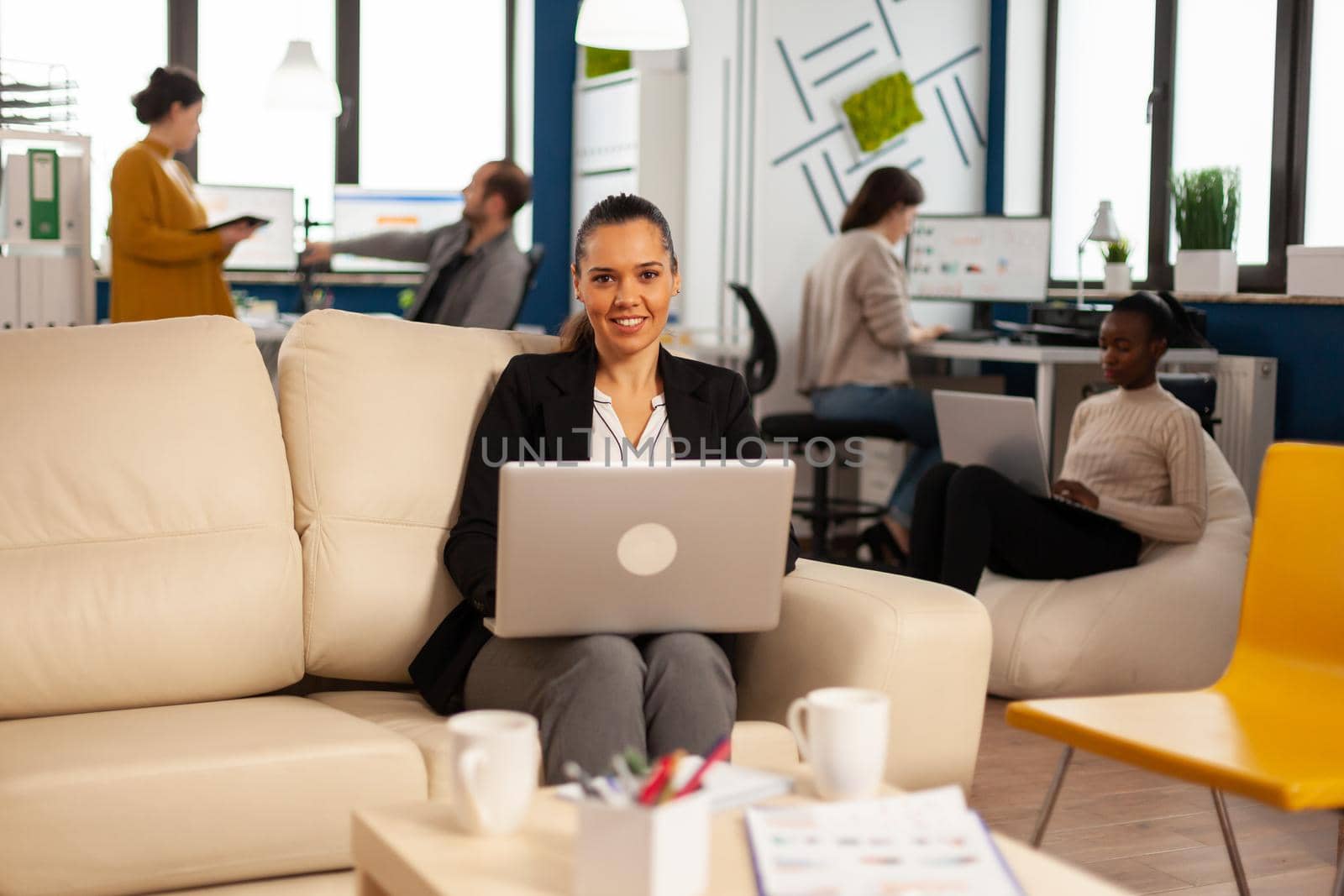 Manager lady writing on laptop looking at camera smiling while diverse colleagues working in background. Multiethnic coworkers talking about startup financial company in modern business office