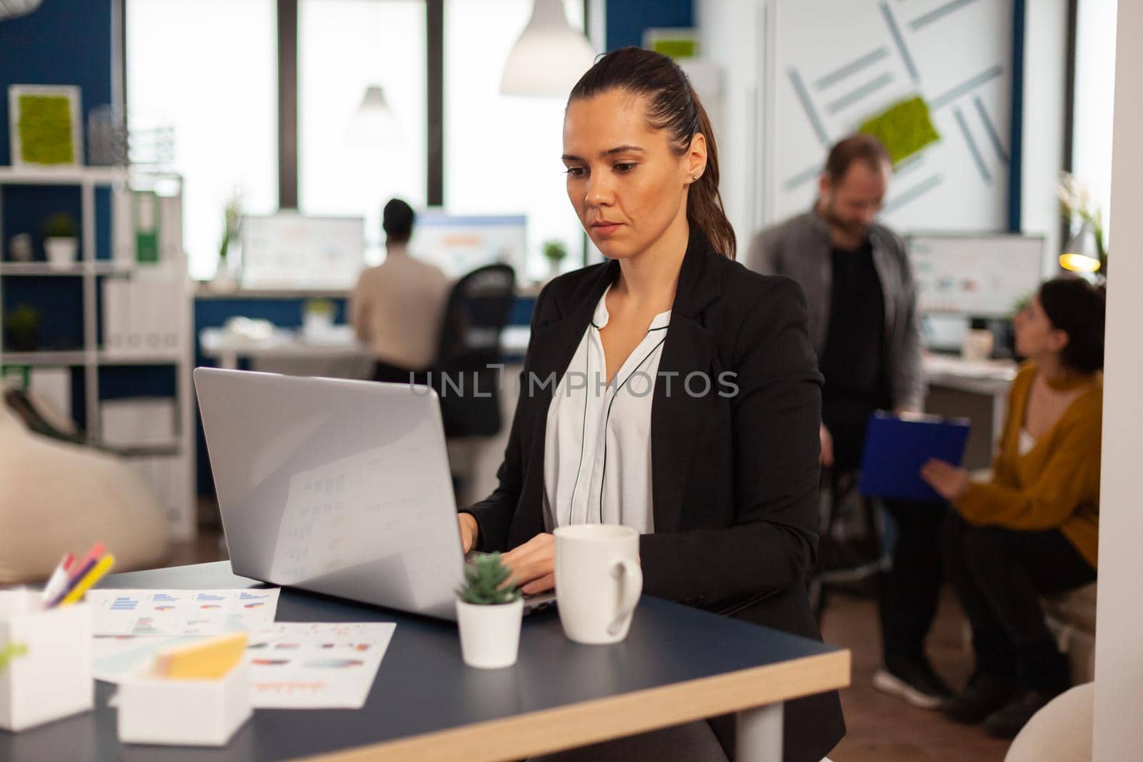 Concentrated business lady writing on laptop computer financial reports sitting at desk in busy start up office enjoying work in creative workplace. Diverse team analyzes statistics data in company