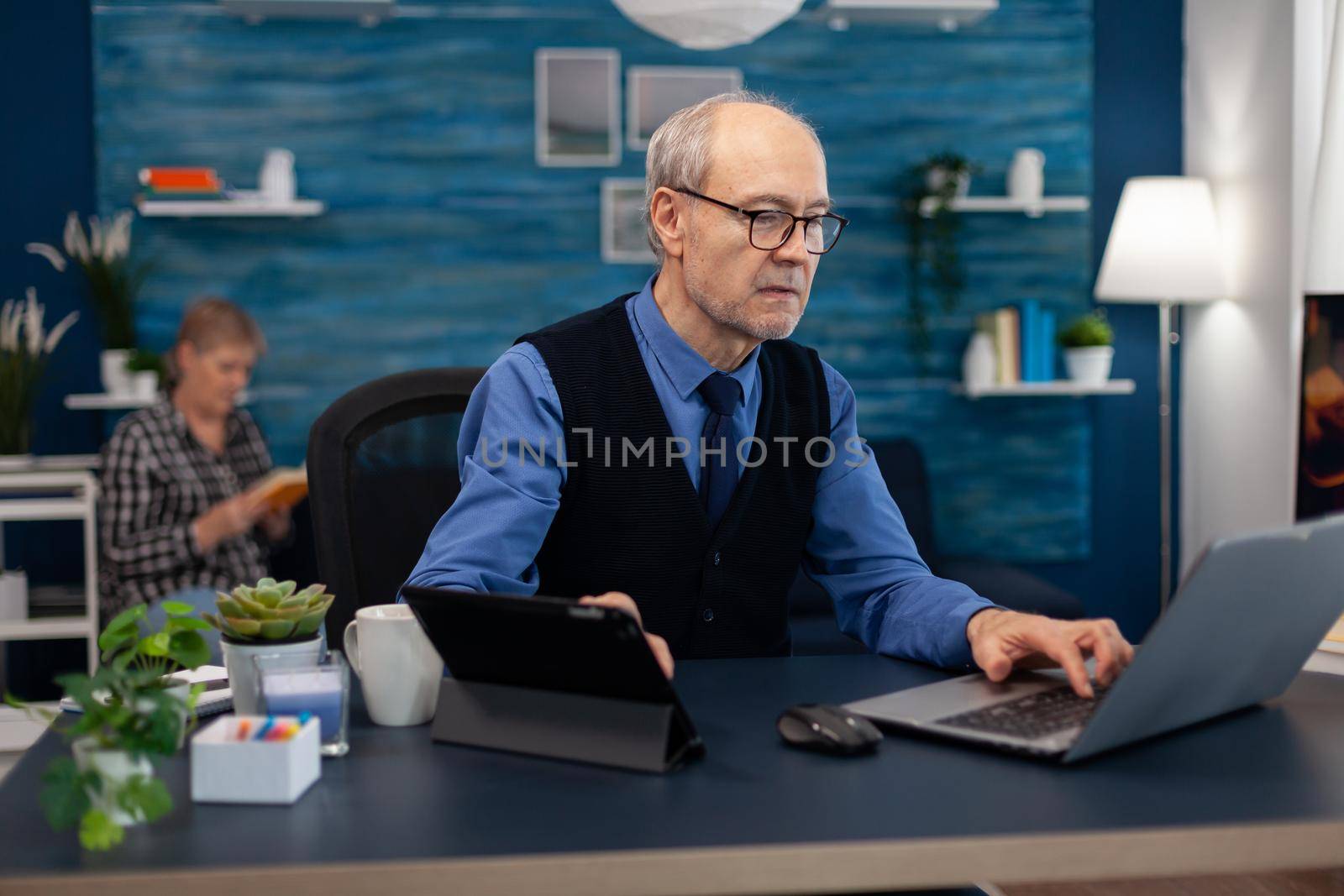 Senior manager working on presentation using laptop and tablet pc sitting at office. Elderly man entrepreneur in home workplace using portable computer sitting at desk while wife is reading a book sitting on sofa.