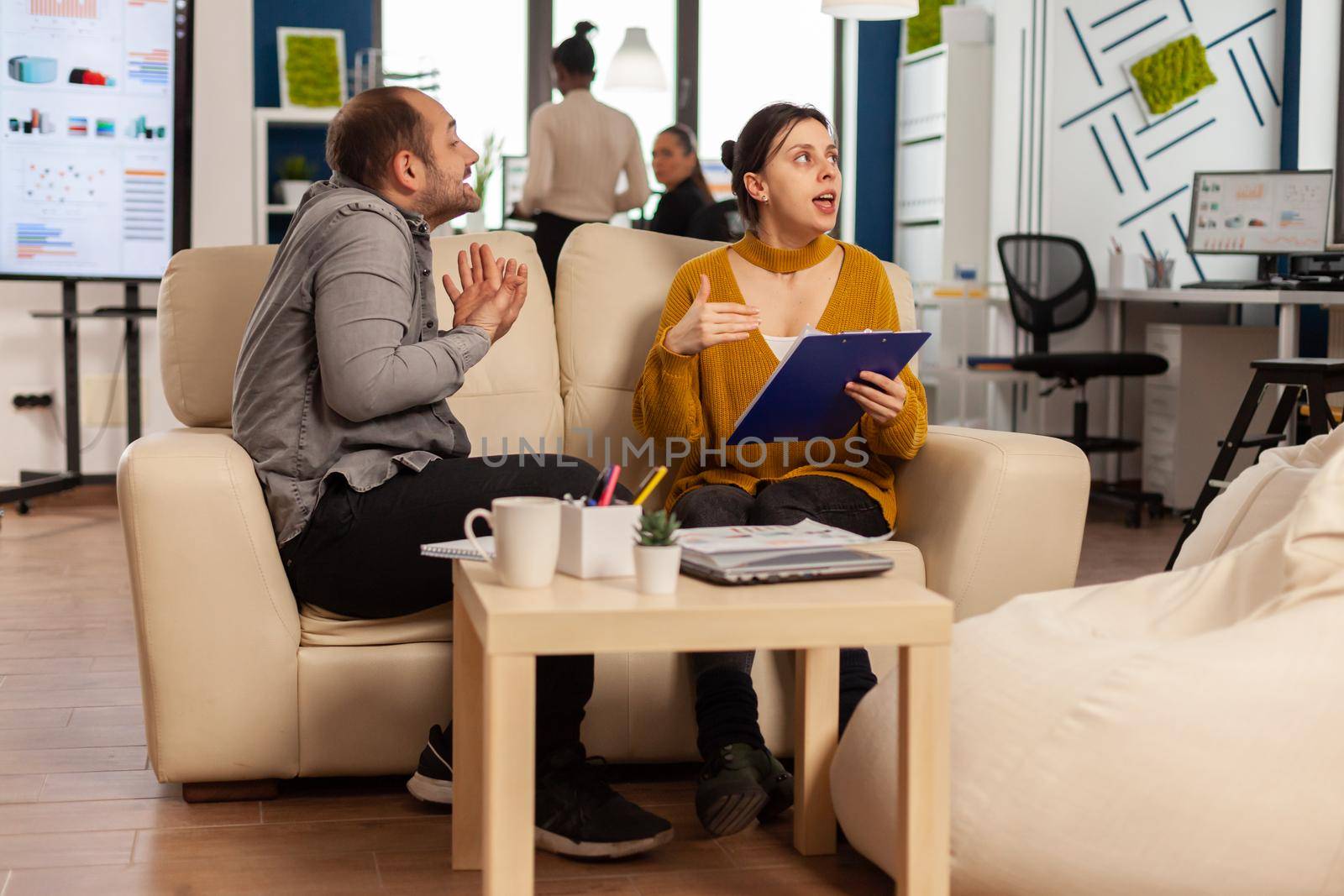Angry man manager arguing woman employee for bad work result, sitting on couch, while diverse colleagues working scared in background. Business woman employee raging about multitasking difficult job.