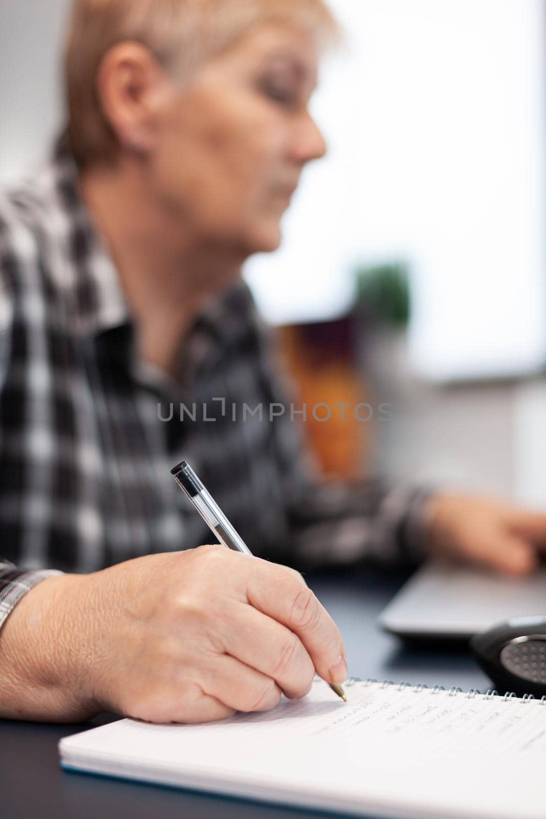 Close up of senior business woman writing notes on notebook. Elderly woman in home living room using moder technoloy laptop for communication sitting at desk indoors.