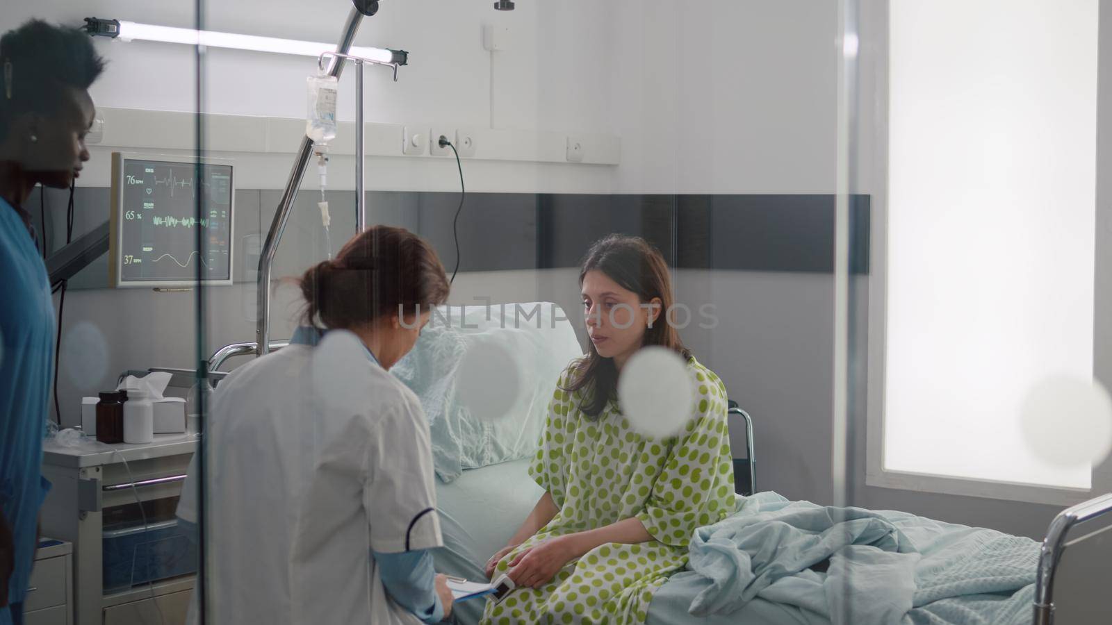 Sick woman talking with doctors while sitting on bed during sickness recovery in hospital ward. Practitioner writing sickness treatment on clipboard examining medical symptom checking heart pulse