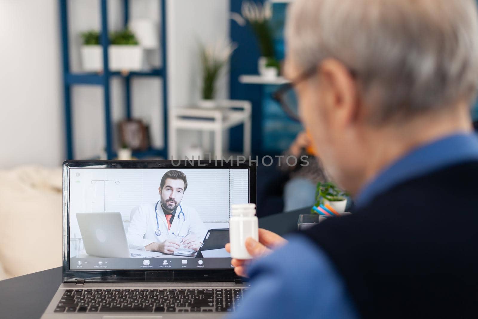 Young doctor talking about treatment with senior man during virtual examination. Elderly man discussing with healthcare practitioner in the course of remote call and wife is reading a book on sofa.