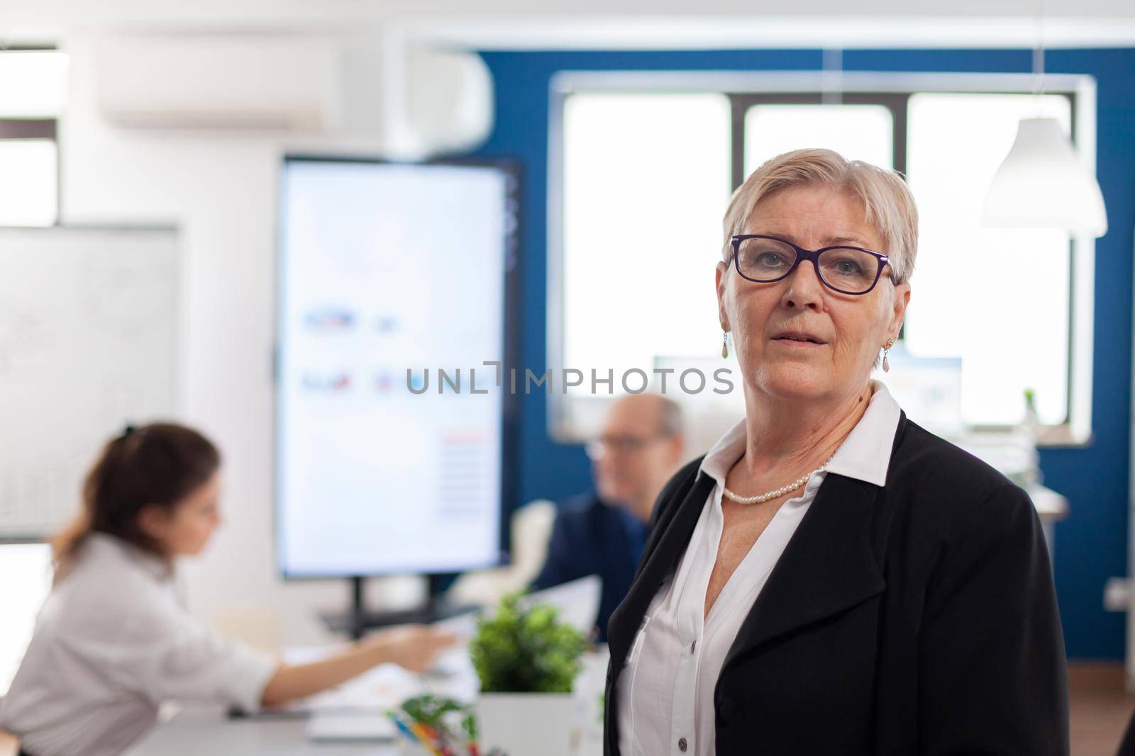 Successful senior entrepreneur standing in front of meeting desk looking at camera. Manager working in professional start up financial business, modern company workplace ready for meeting.