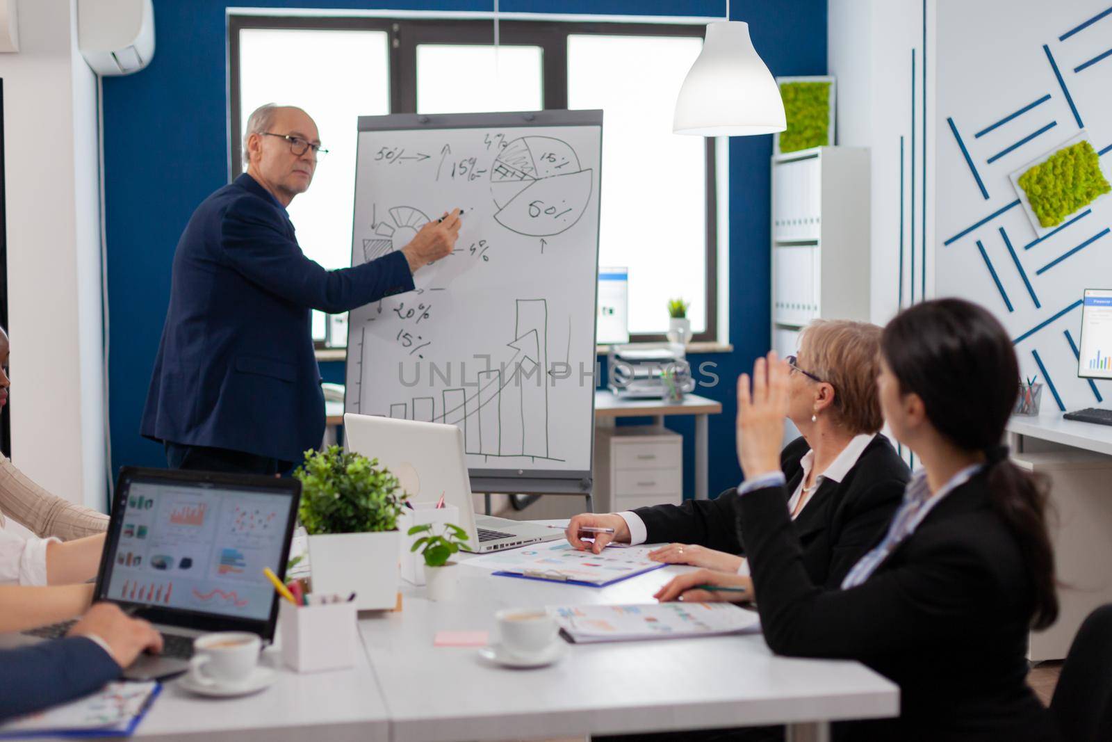 Senior businessman in eyeglasses and formal suit drawing financial plan on flipchart. Serious speaker boss executive, business trainer explaining development strategy to motivated mixed race employees.