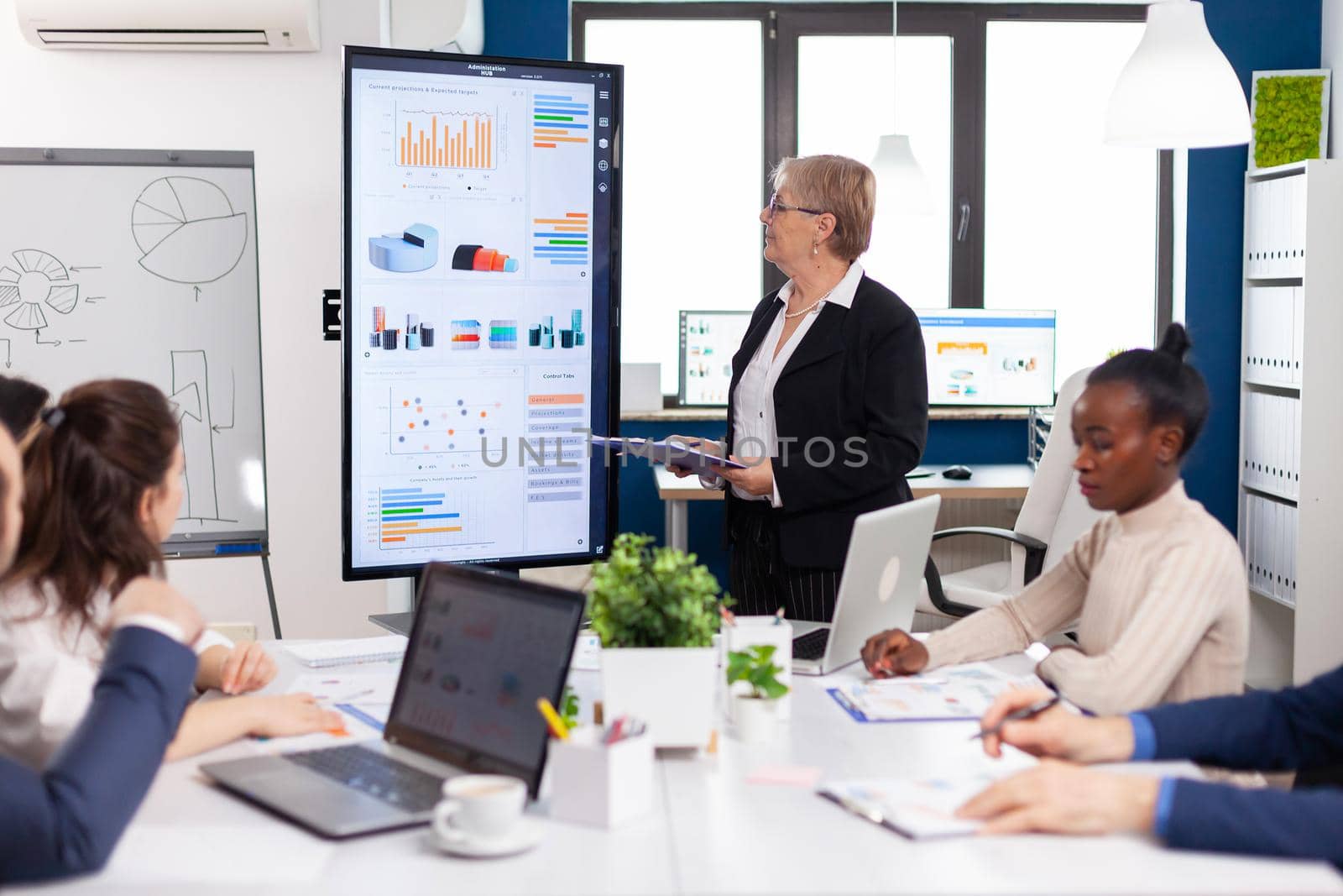 Elderly project manager pointing at desktop presenting statistical data, briefing diverse group of employees. Multiethnical businesspeople working in professional startup financial office during conference meeting