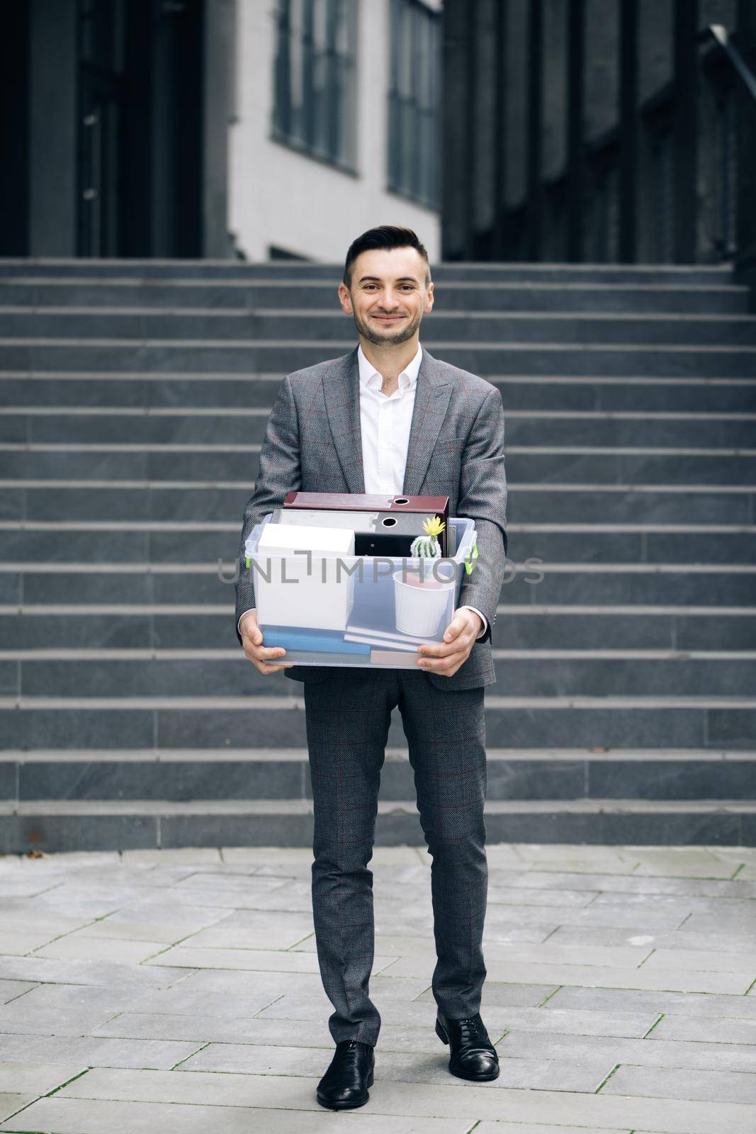 Young happy businessman returns to work after vacation. New employee on first day at work carrying box with stuff to his new workplace. Concept of work career and success by uflypro