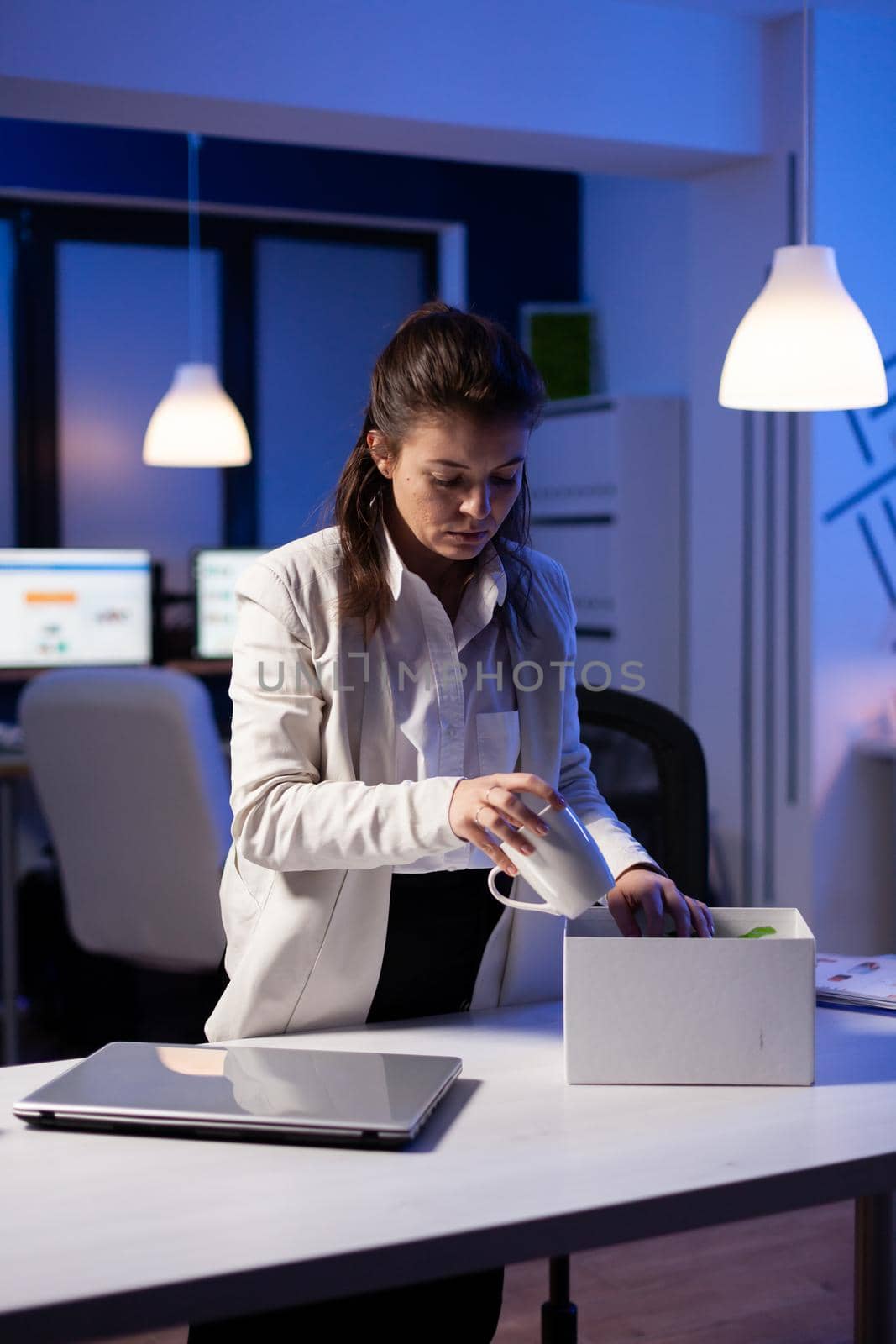 Depressed dismissed employee putting her stuff in white box from table at office as being fired. Unemployed packing things late at night. woman leaving workplace office in midnight