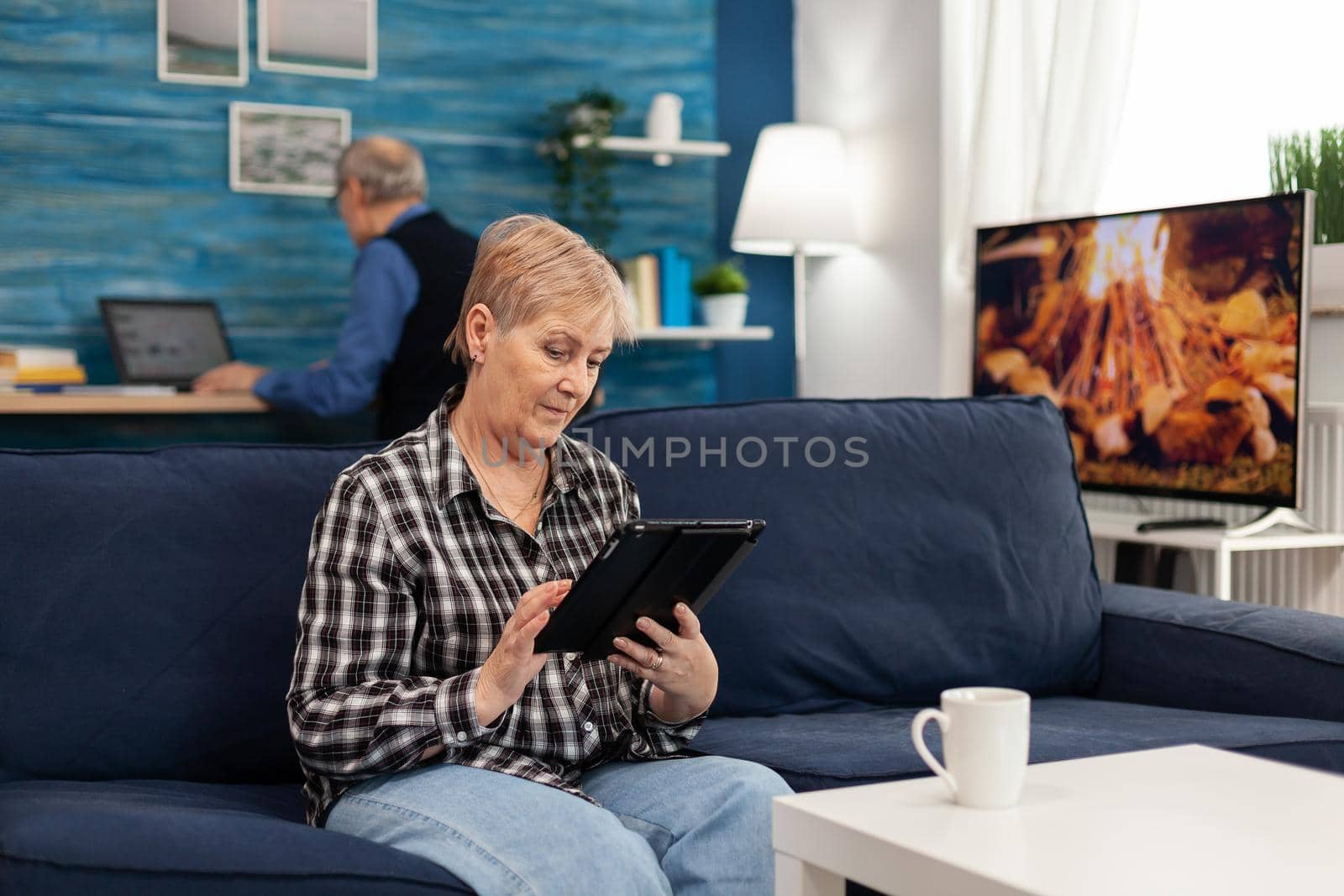 Senior lady scrolling on digital device relaxing on sofa. Elderly woman using moder technoloy tablet pc in home living room and husband working on laptop computer