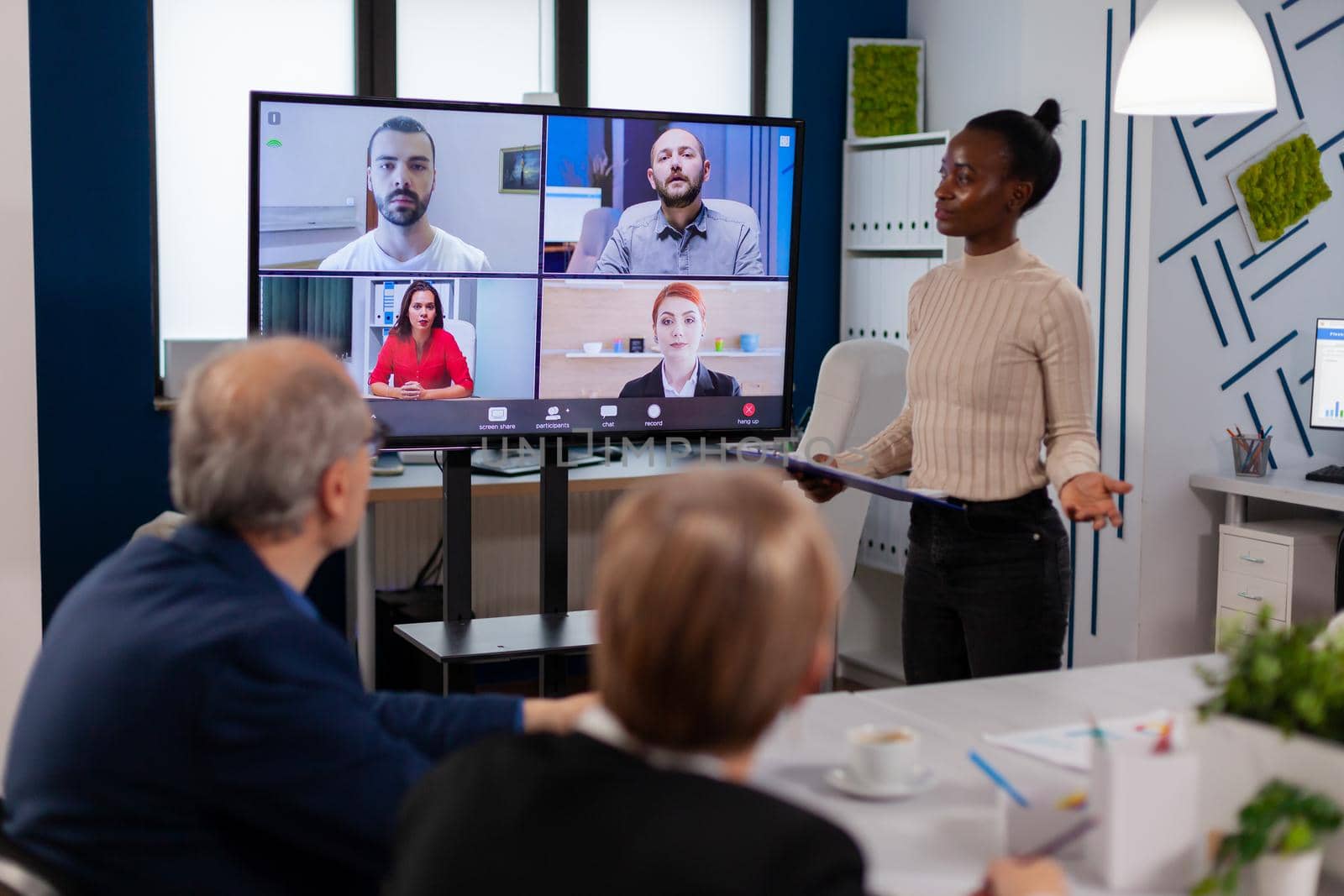 Black manager woman talking with remotely colleagues on video call at tv screen, presenting new business partners. Business people talking to webcam, do online conference participate internet brainstorming, distance office discussion.
