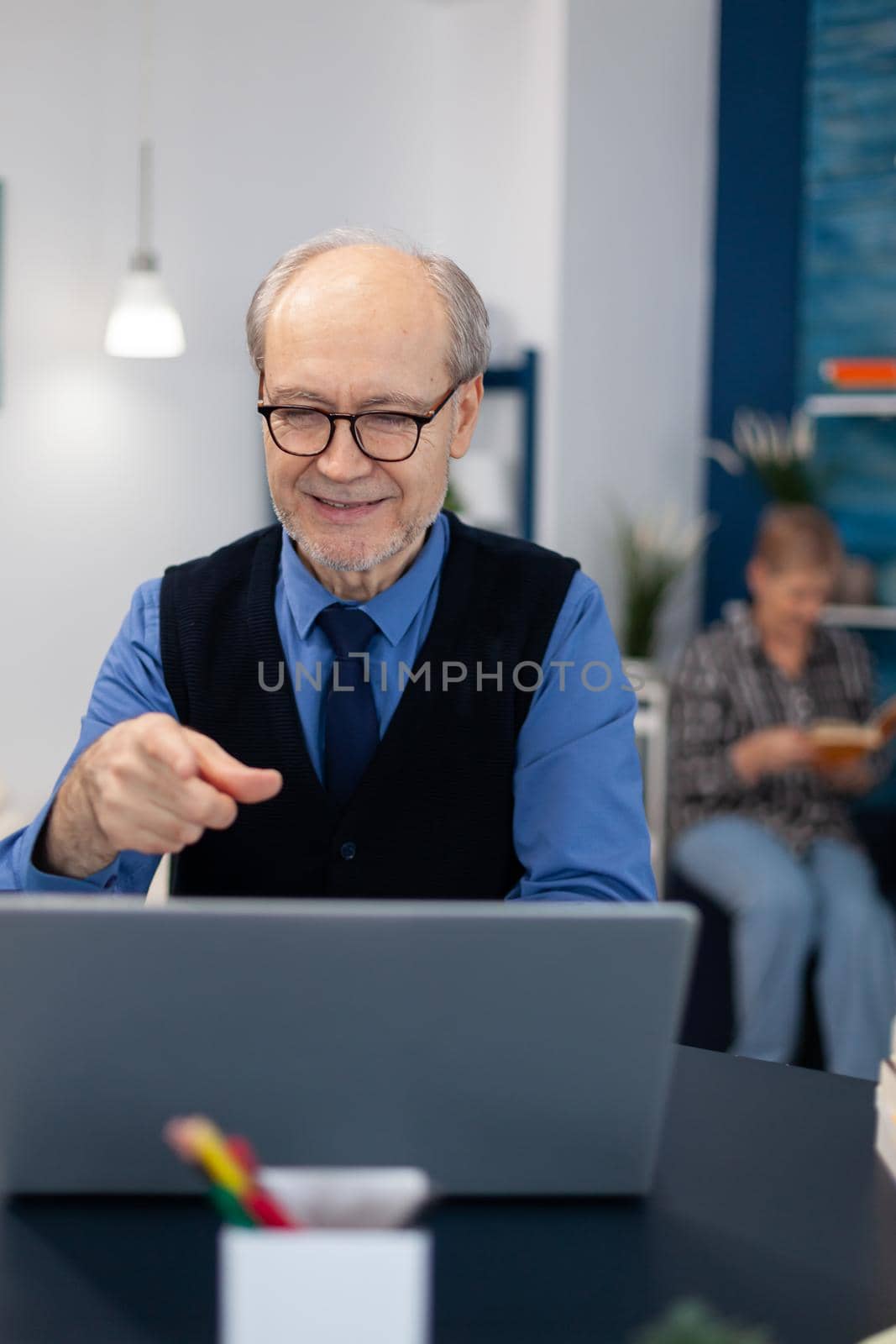 Happy senior man pointing at laptop during video conference. Cheerful pensioner with gray hair smiling at laptop webcam while chatting sitting at desk .