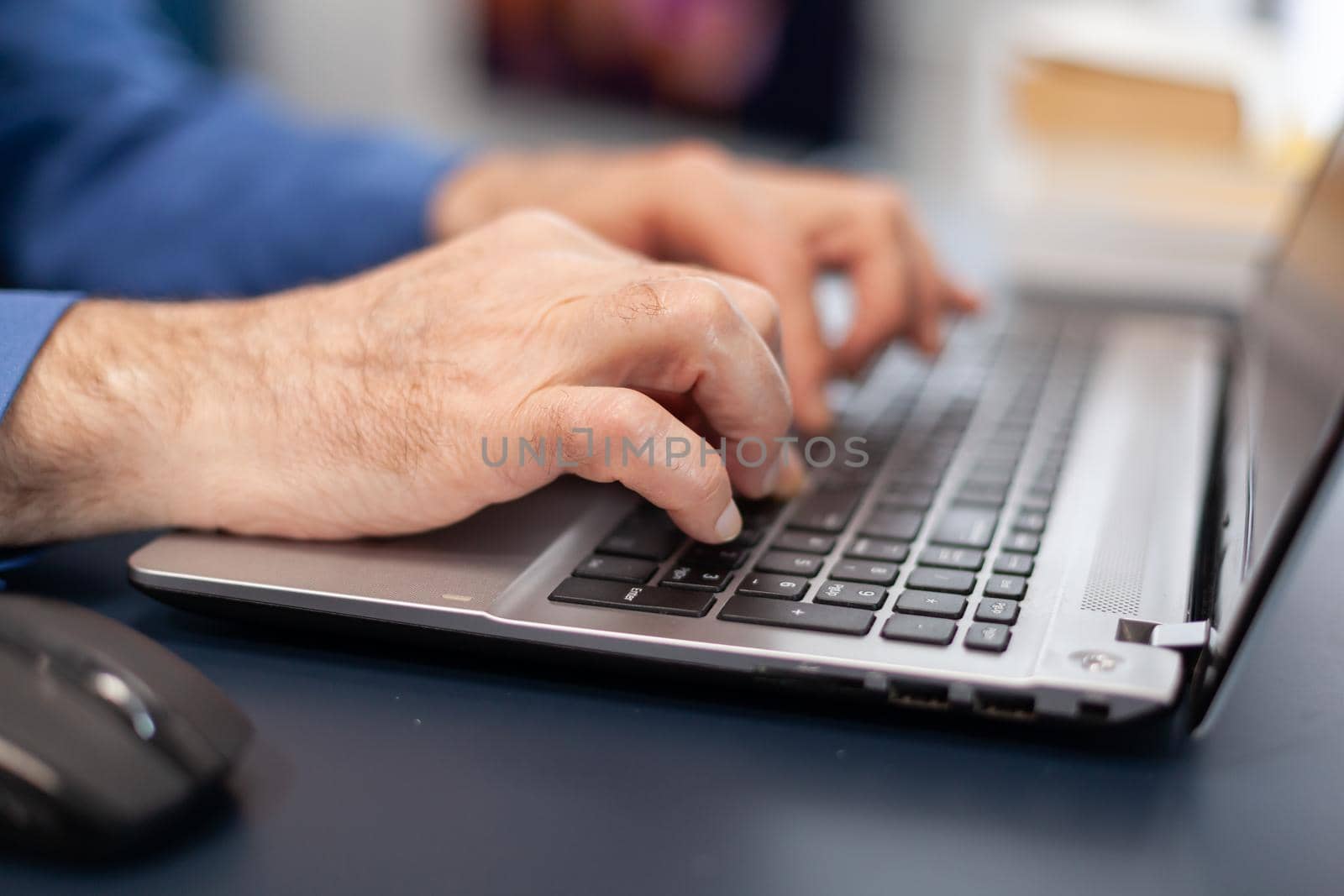 Close up of senior man hands typing on laptop keyboard. Elderly man entrepreneur in home workplace using portable computer sitting at desk while wife is holding tv remote