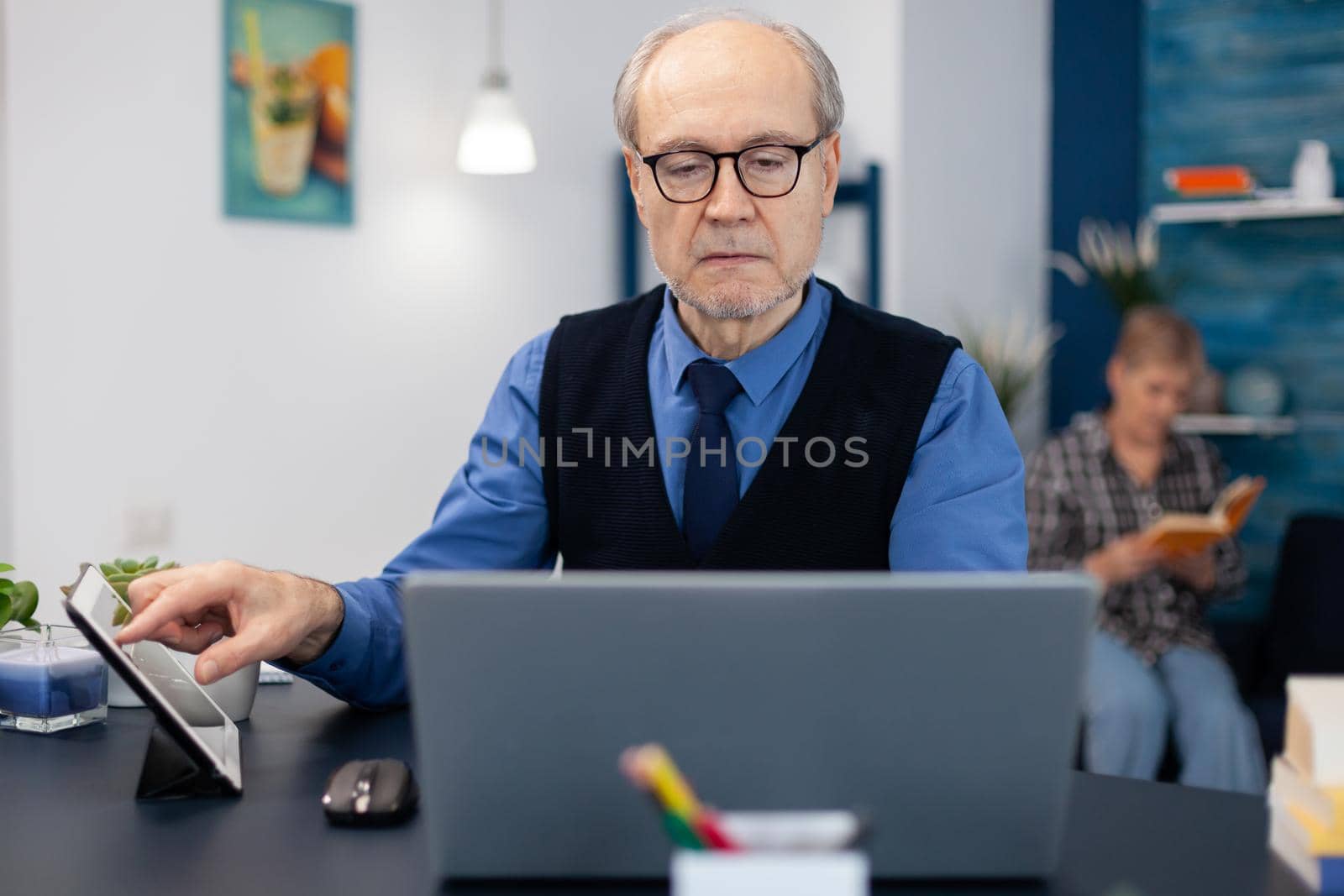 Portrait of mature man doing websurfing on laptop from home office. Elderly man entrepreneur in home workplace using portable computer sitting at desk while wife is reading a book sitting on sofa.
