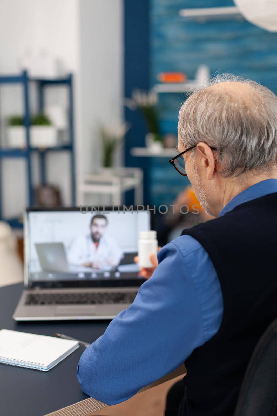 Senior man showing pills bottle to doctor while using laptop for telemedicine. Elderly man discussing with healthcare practitioner in the course of remote call and wife is reading a book on sofa.