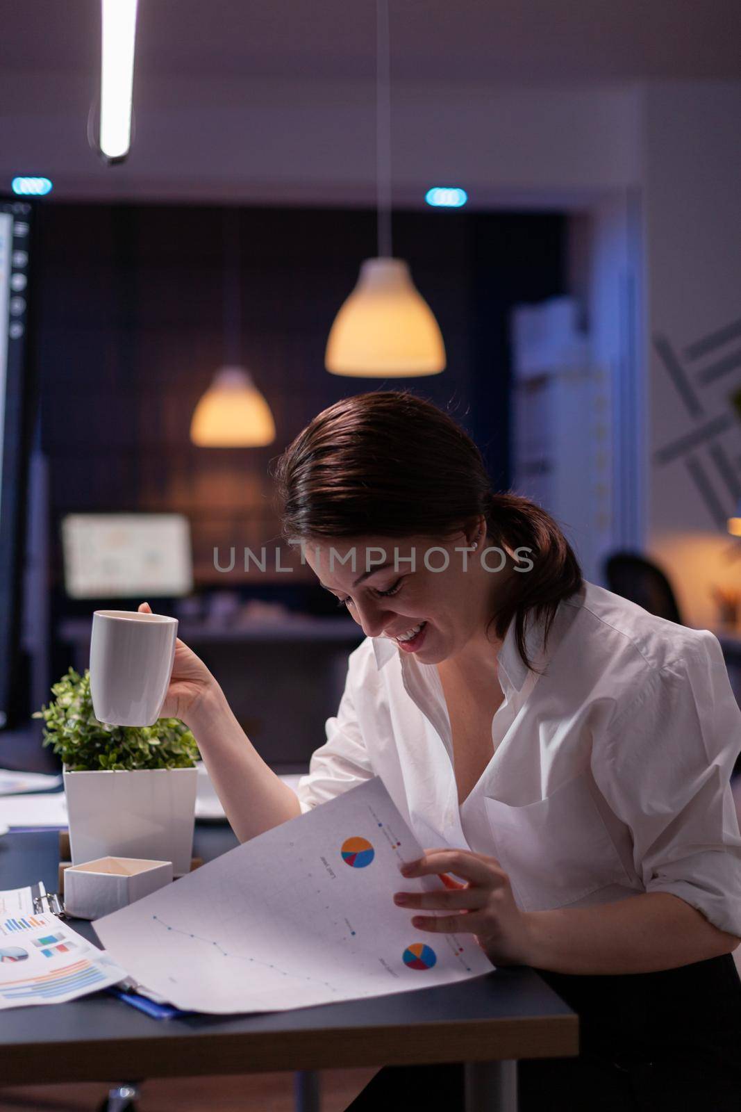 Workaholic businesswoman looking at reports presentation on monitor comparing with financial chart on paperwork. Focused manager working in business company meeting office room late at night