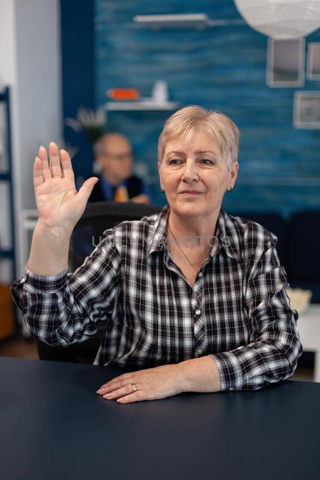 Thoughtfull elderly lady greeting people in the course of online conference. Happy senior woman with gray hair saying hello at camera during video call in home living room.