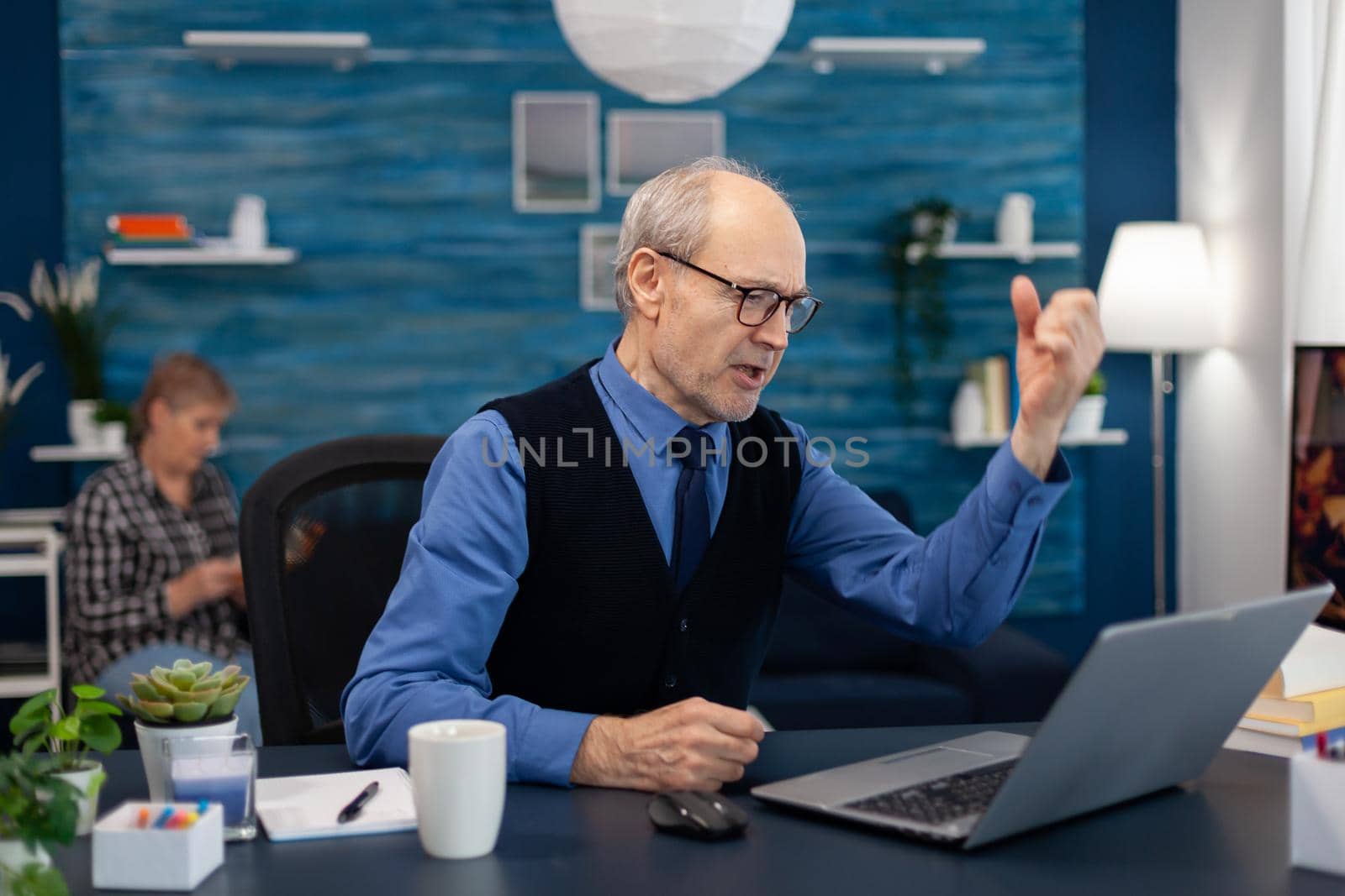 Excited senior man celebrating good news while working on laptop from home office. Elderly man entrepreneur in home workplace using portable computer sitting at desk while wife is reading a book sitting on sofa.
