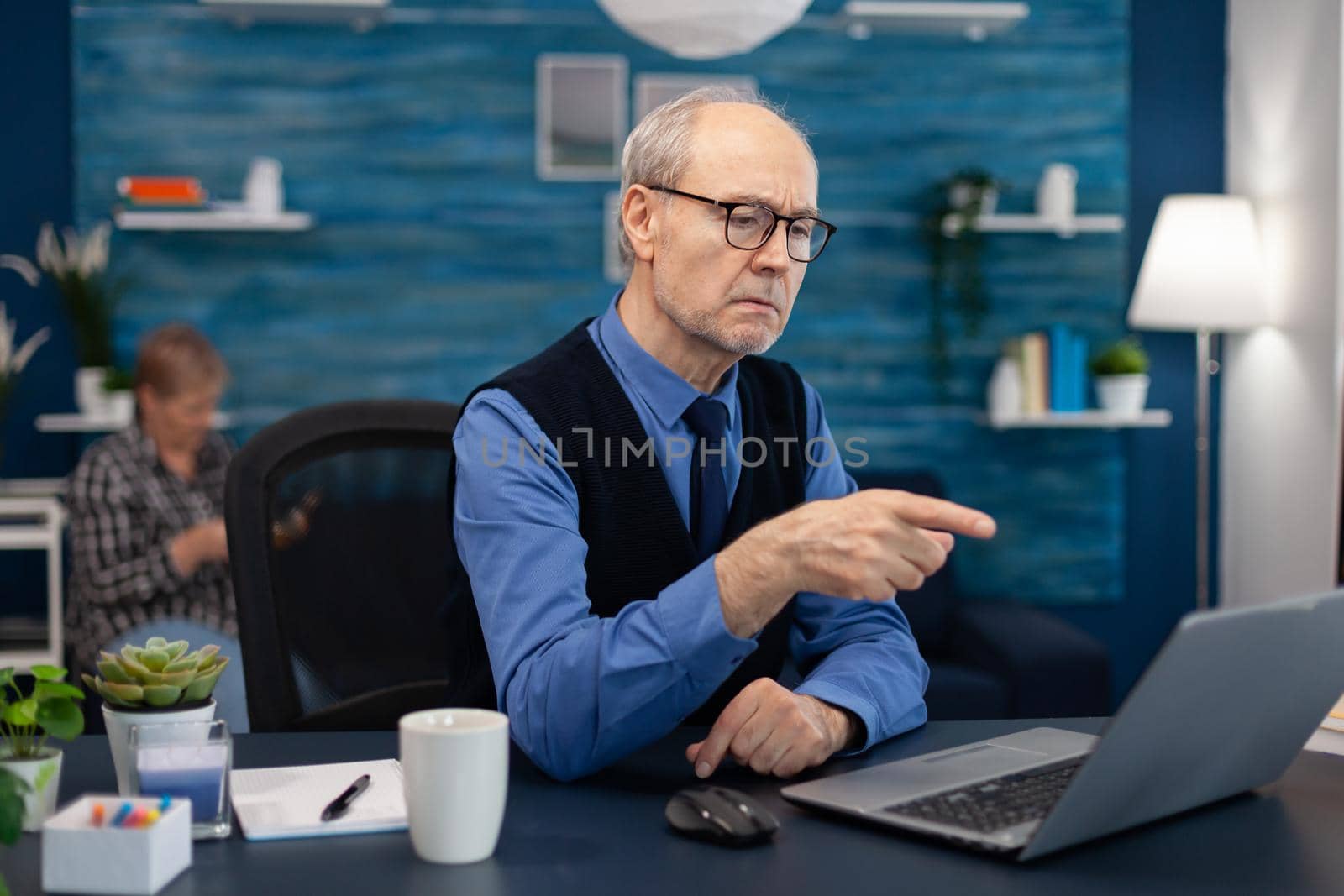 Thoughtfull senior businessman pointing at laptop whille working from home. Elderly man entrepreneur in home workplace using portable computer sitting at desk while wife is holding tv remote.