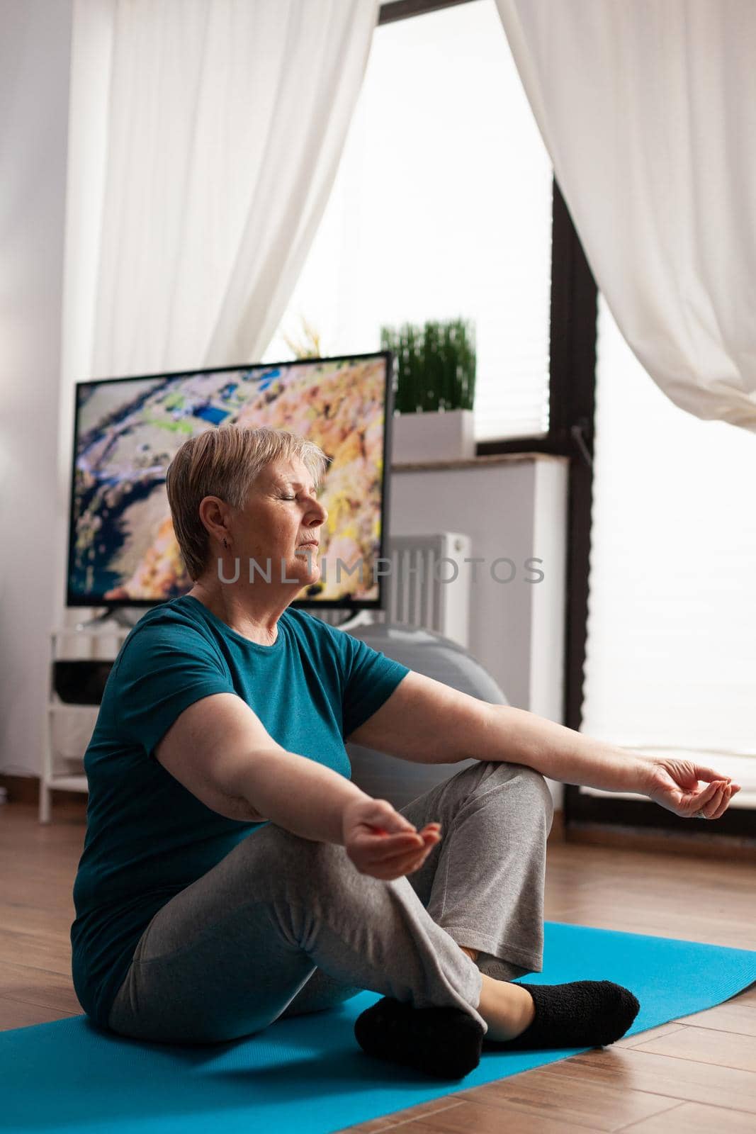 Peaceful senior woman sitting comfortable in lotus position on yoga mat with closed eyes by DCStudio