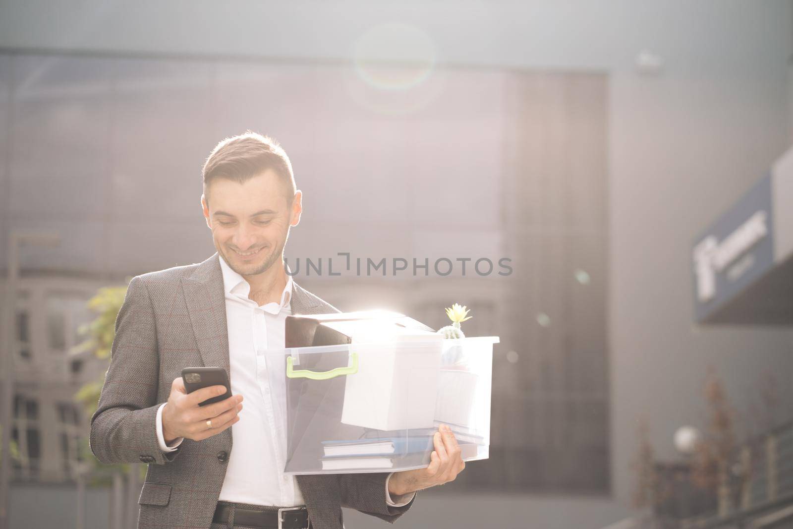 Happy businessman stand smiling use phone near business center. Businessman with box of personal stuff uses phone texting scrolling tapping. Portrait suit career male office handsome technology.