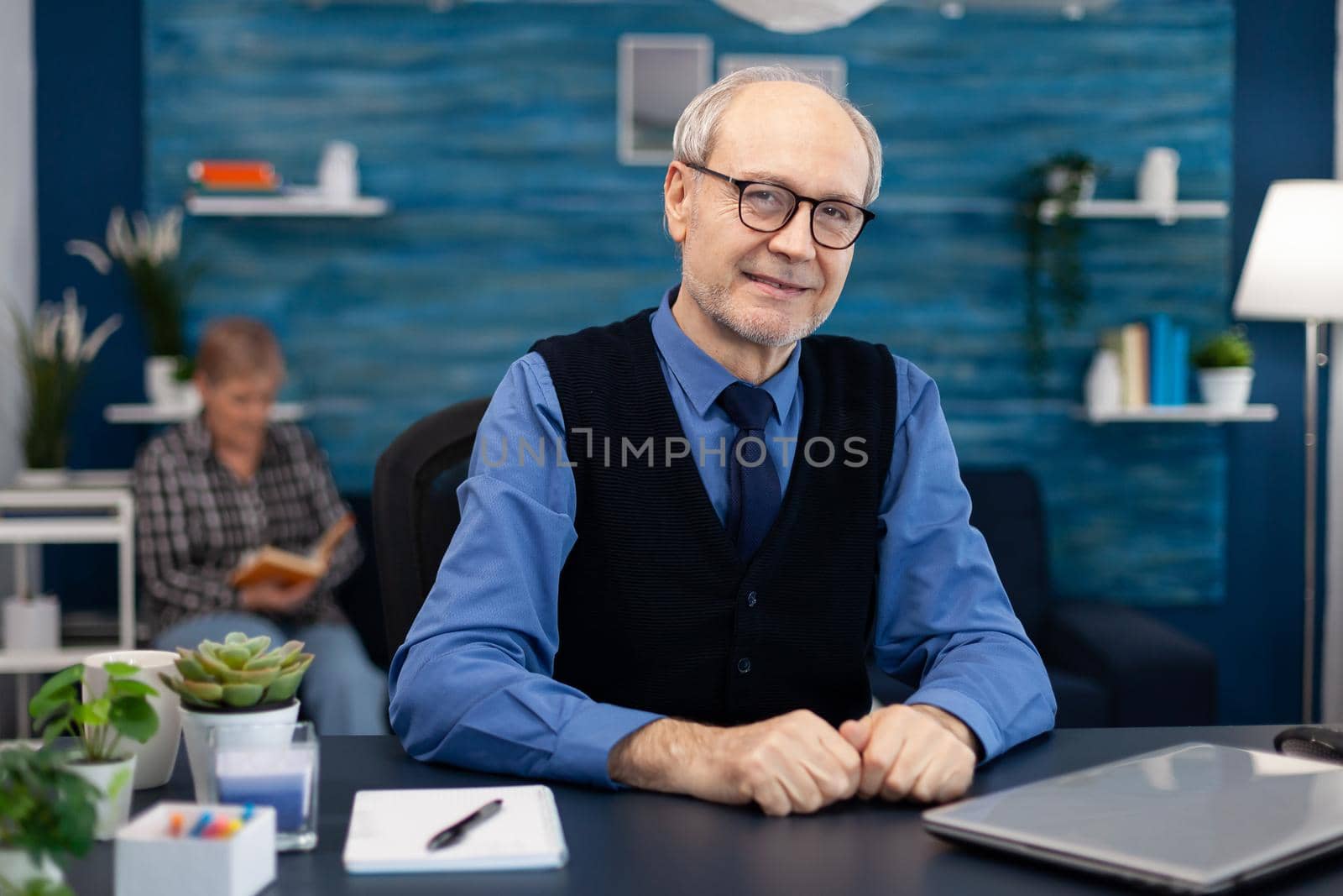 Portrait of senior man sitting at desk office looking at camera. Elderly man entrepreneur in home workplace using portable computer sitting at desk while wife is reading a book sitting on sofa.
