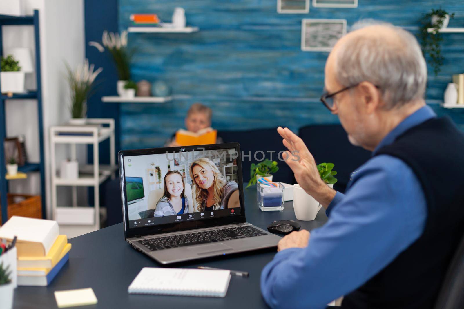 Elderly man waving at camera during online call with his niece. Happy grandfather saying hello in the course of online video confencere with family from living room.
