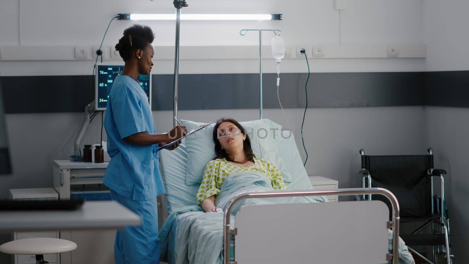 Afro american asisstant checking sick woman while sitting in bed in hospital ward by DCStudio