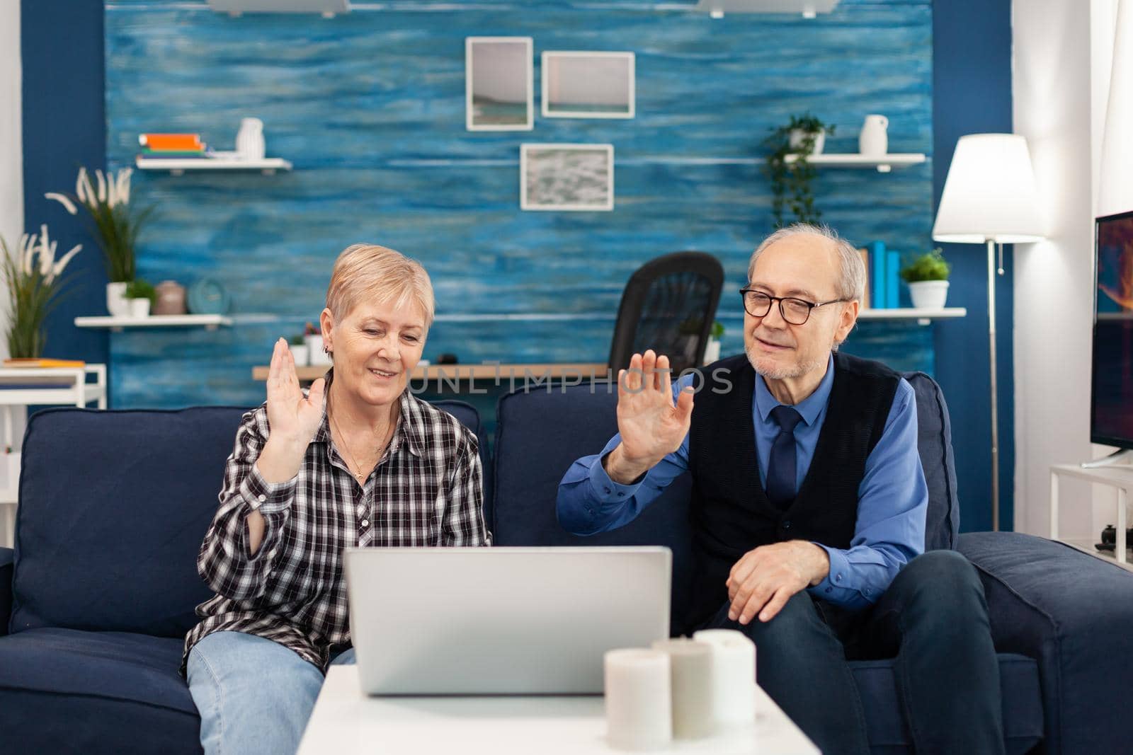 Cheerful senior man and woman waving during online call with family. Happy elderly man and woman saying hello to laptop webcam sitting on sofa in the course of video conference.