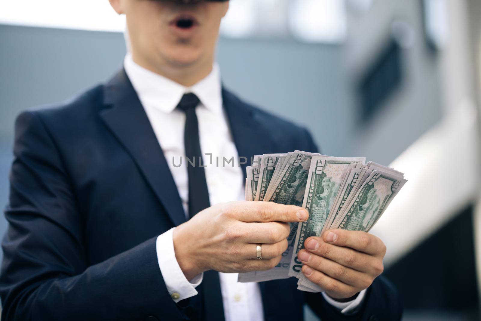 Rich man wearing VR headset counting money and smiling. Young businessman standing with pack of dollars. Richness and success concept. New technology offers new 3D dimensions. by uflypro