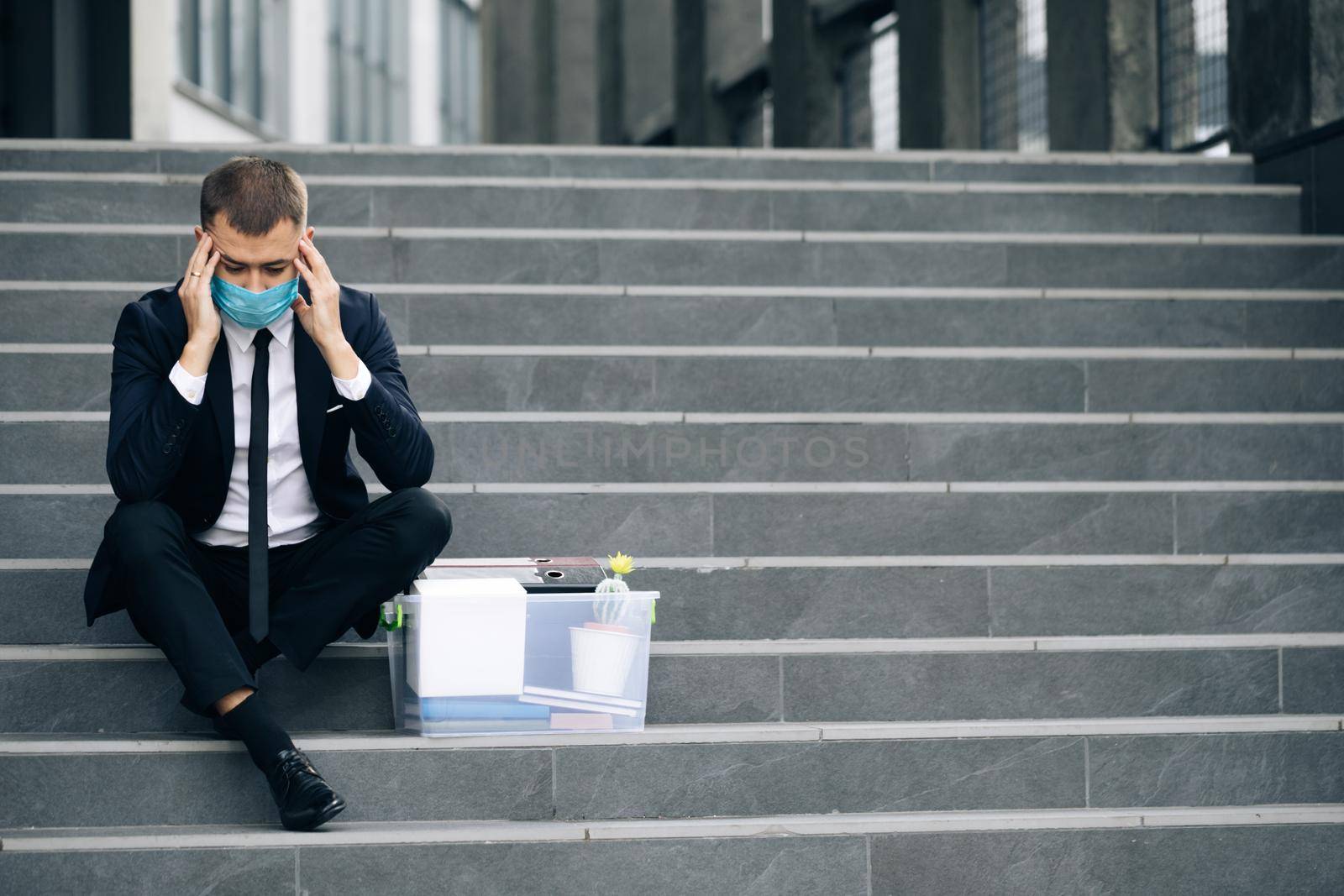 Fired male office worker in medical mask sitting on stairs in depression with box of stuff. Anxious concept. Workless man in despair. Unemployed businessman lost his business by uflypro