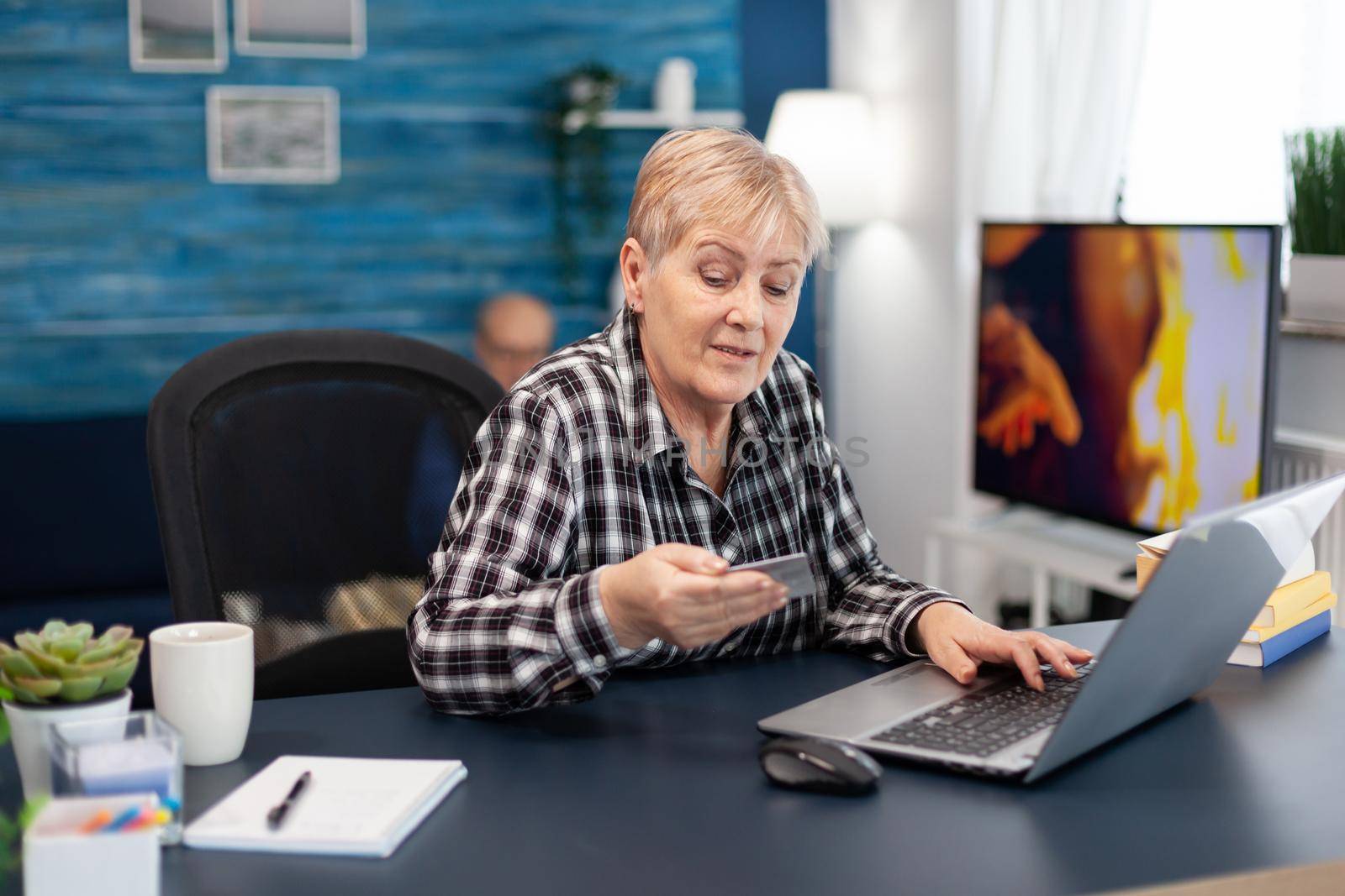 Senior woman reading cvv code from credit card sitting in front of laptop. Joyful elderly woman using online banking for payment transcation surfing on internet from home living room.