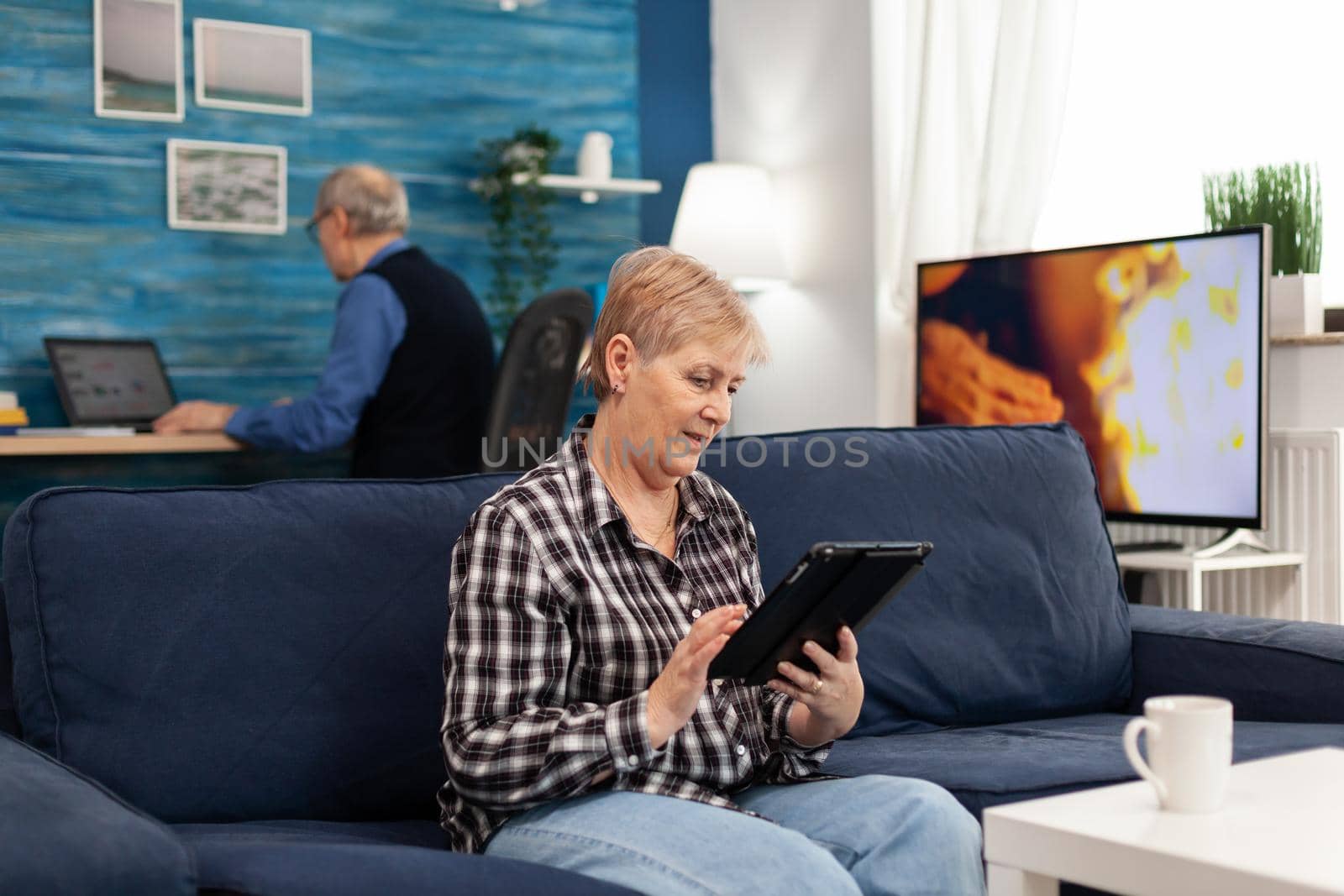 Happy mature woman relaxing on couch enjoying retirement lifestyle. Elderly woman using moder technoloy tablet pc in home living room and husband working on laptop computer
