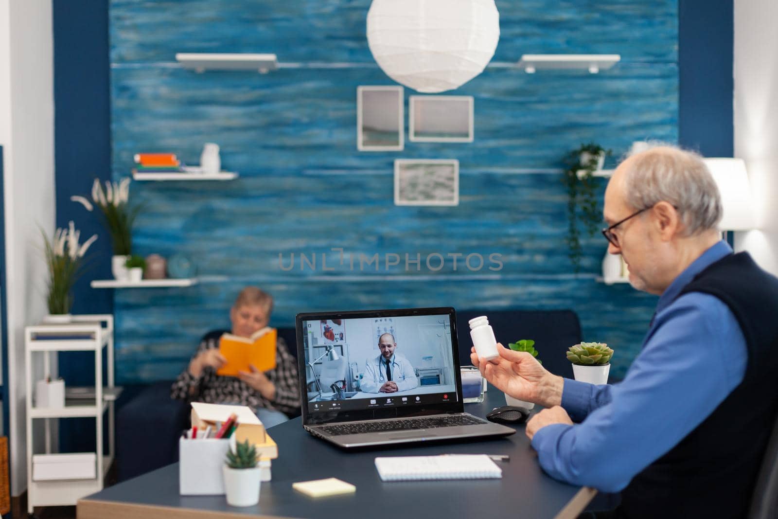 Senior man listening doctor during video conference holding pills bottle. Elderly man discussing with healthcare practitioner in the course of remote call and wife is reading a book on sofa.