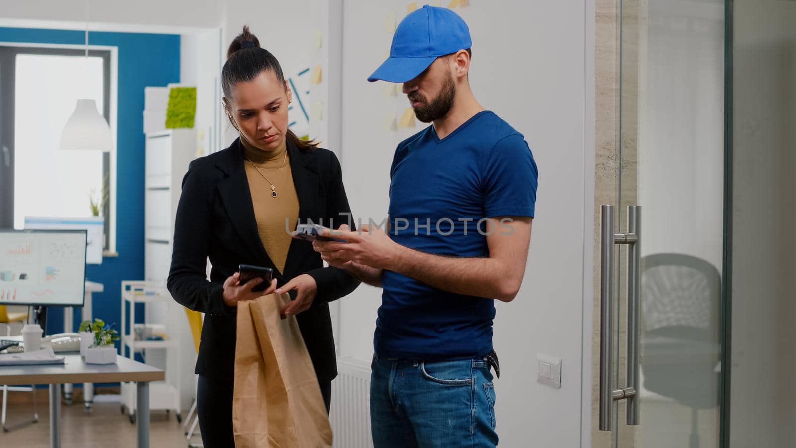 Delivery man bringing takeaway food meal order to businesswoman in startup company office during takeout lunchtime. Entrepreneur receiving lunch paying with smartphone using POS contactless service