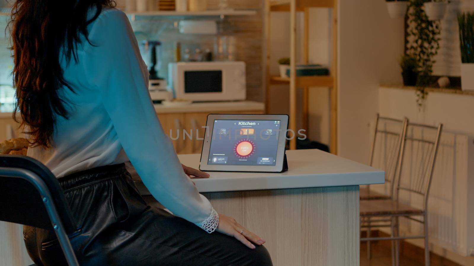 Woman looking at tablet in house with automation lighting system, sitting in kitchen switching on bulbs using voice command. Person controlling lights using wireless gadget with smart home application