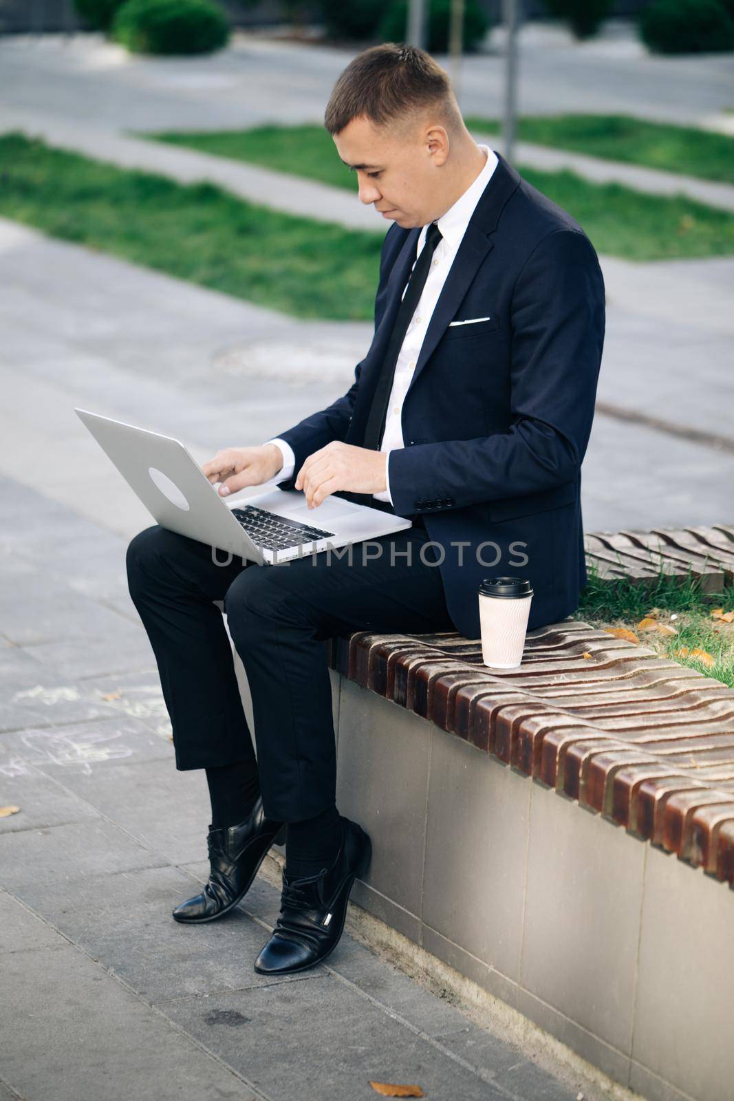 Distance working. Isolated man in a suit. Handsome young businessman sitting outdoors working with his laptop. Crisis 2020. Coronavirus outbreak.