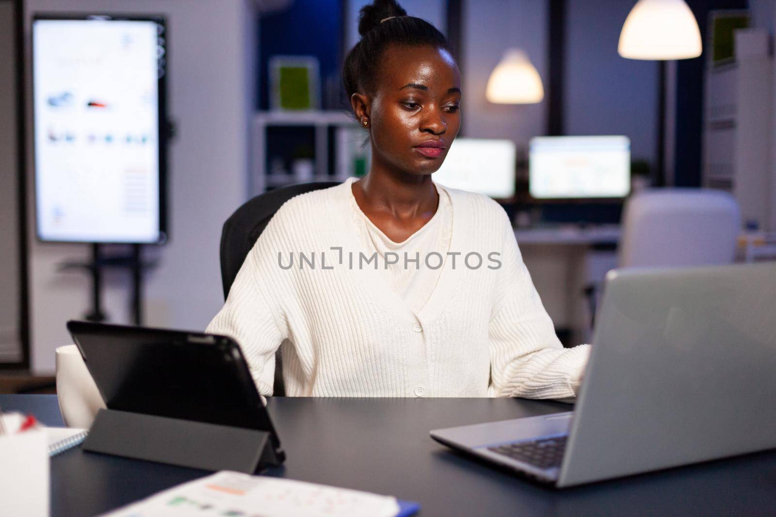 Workaholic african manager trying to finish deadline checking charts on tablet pc and laptop. Busy multitasking employee analysing financial statistics overworking writing, searching.