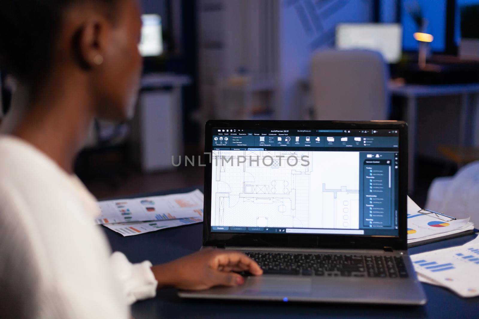 Over shoulder view of african architect working late at night in office using cad software, house blue prints. Industrial woman employee studying prototype idea showing cad software on device display.